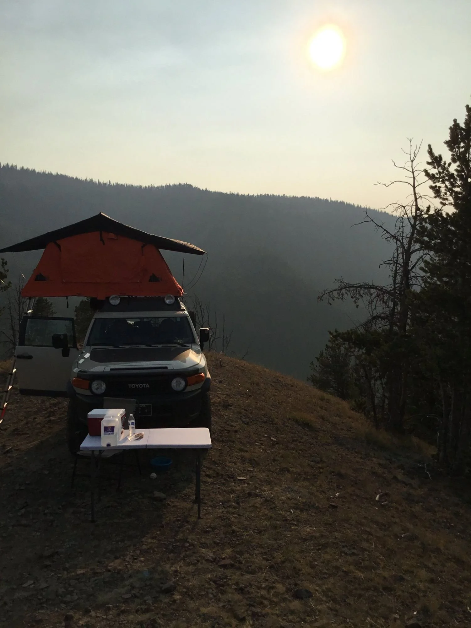 Hazy orange sun over a smoky valley with an FJ Cruiser and rooftop tent on the ridge on Little Bear Road in the Gallatin Range near Bozeman, Montana
