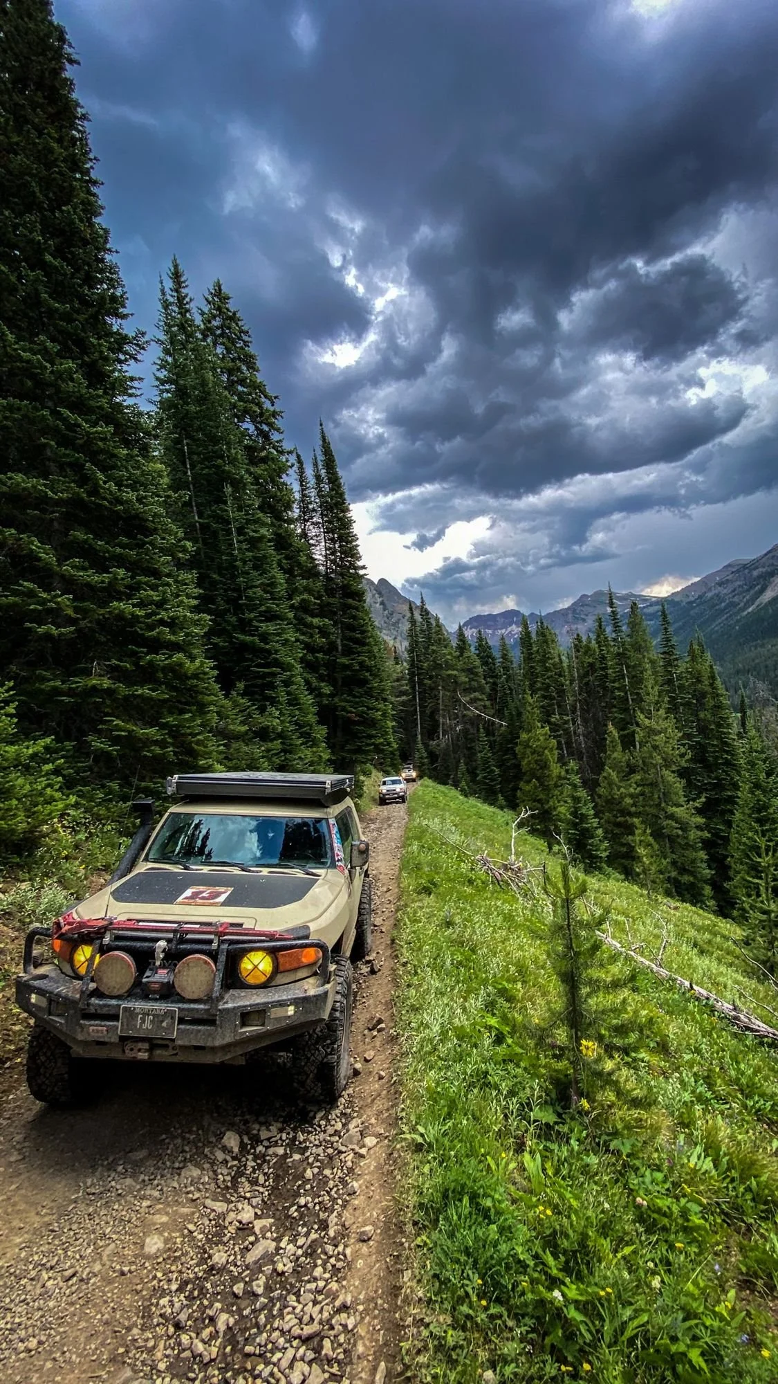 Tan Toyota FJ Cruiser on a narrow hillside road under dark storm clouds on the Independence road above the Boulder River, Montana