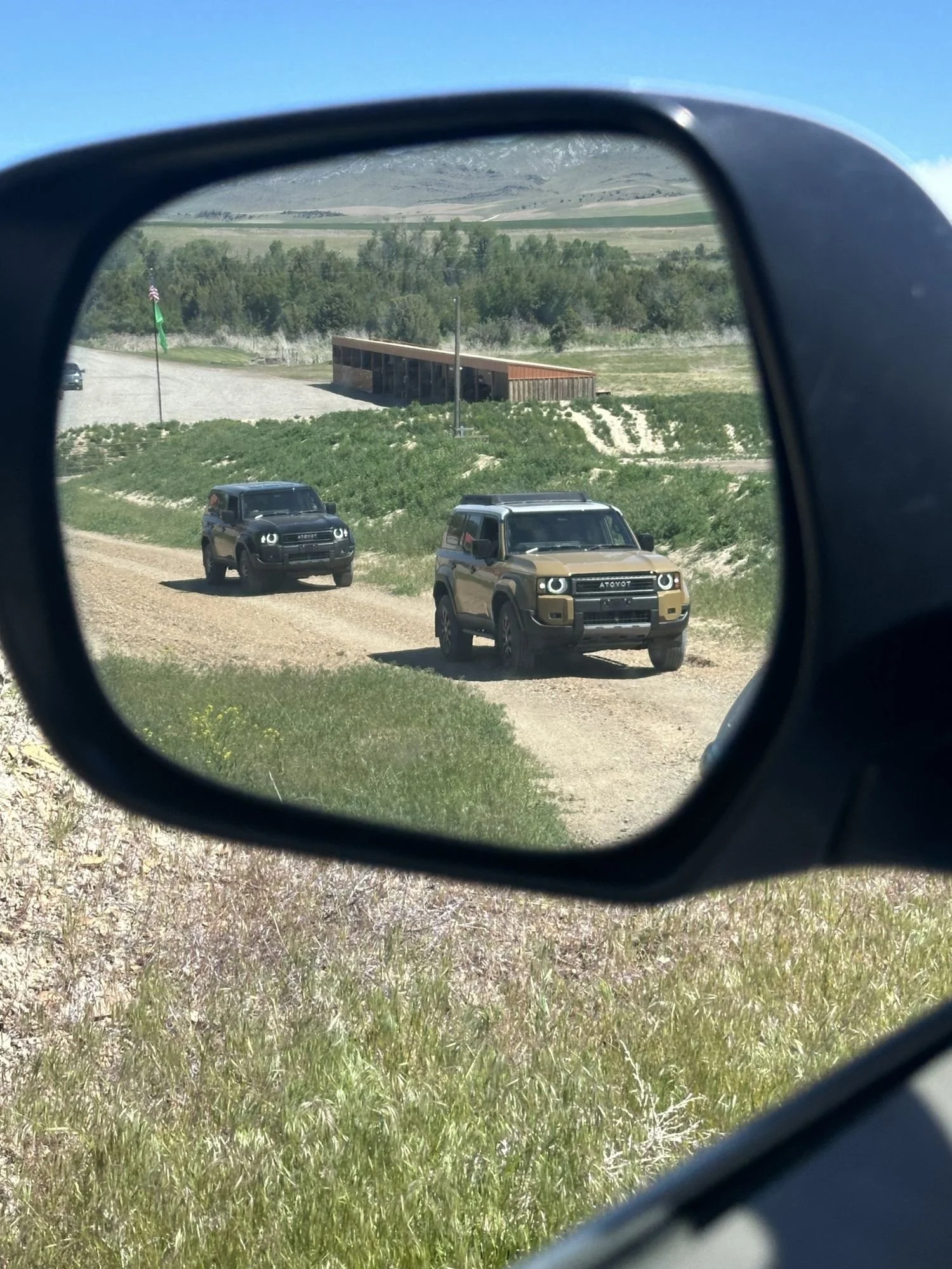Side mirror reflecting a black and a tan Land Cruiser 250 on a grassy course at XOverland Ranch near Bozeman, Montana