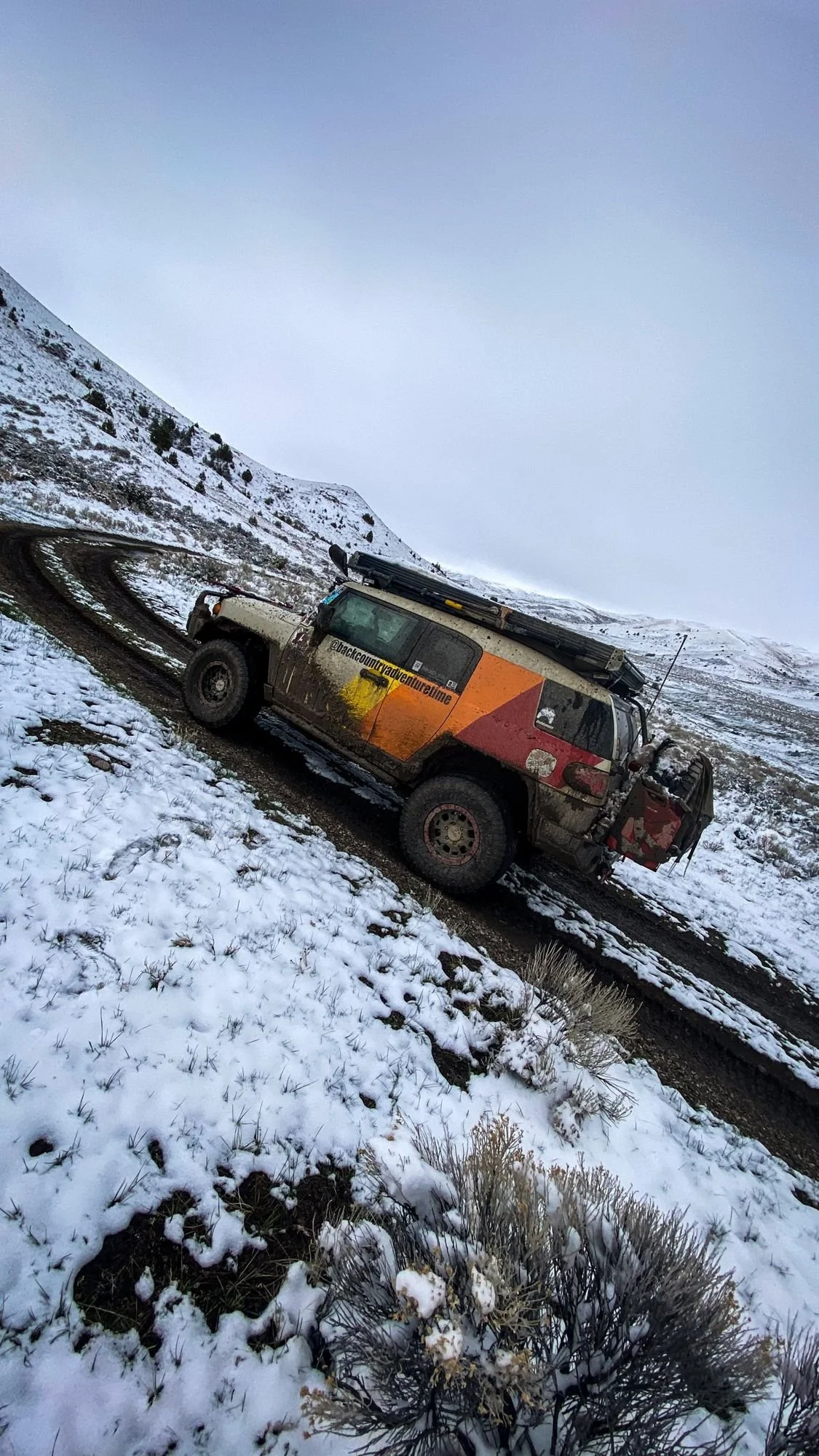 Toyota FJ Cruiser with orange and red graphics tilted on an off-camber snowy hillside road on a 406 Cruisers run in the upper Ruby River country, Montana