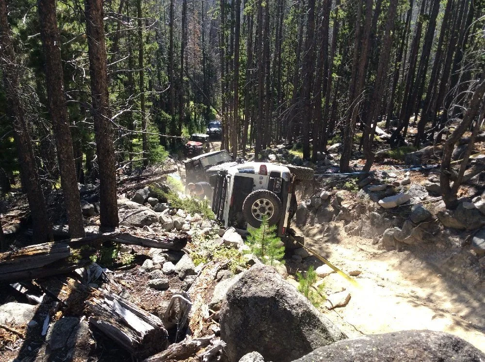 View down a steep rocky trail to a white FJ Cruiser on its side in the trees on a rocky forest trail in Montana