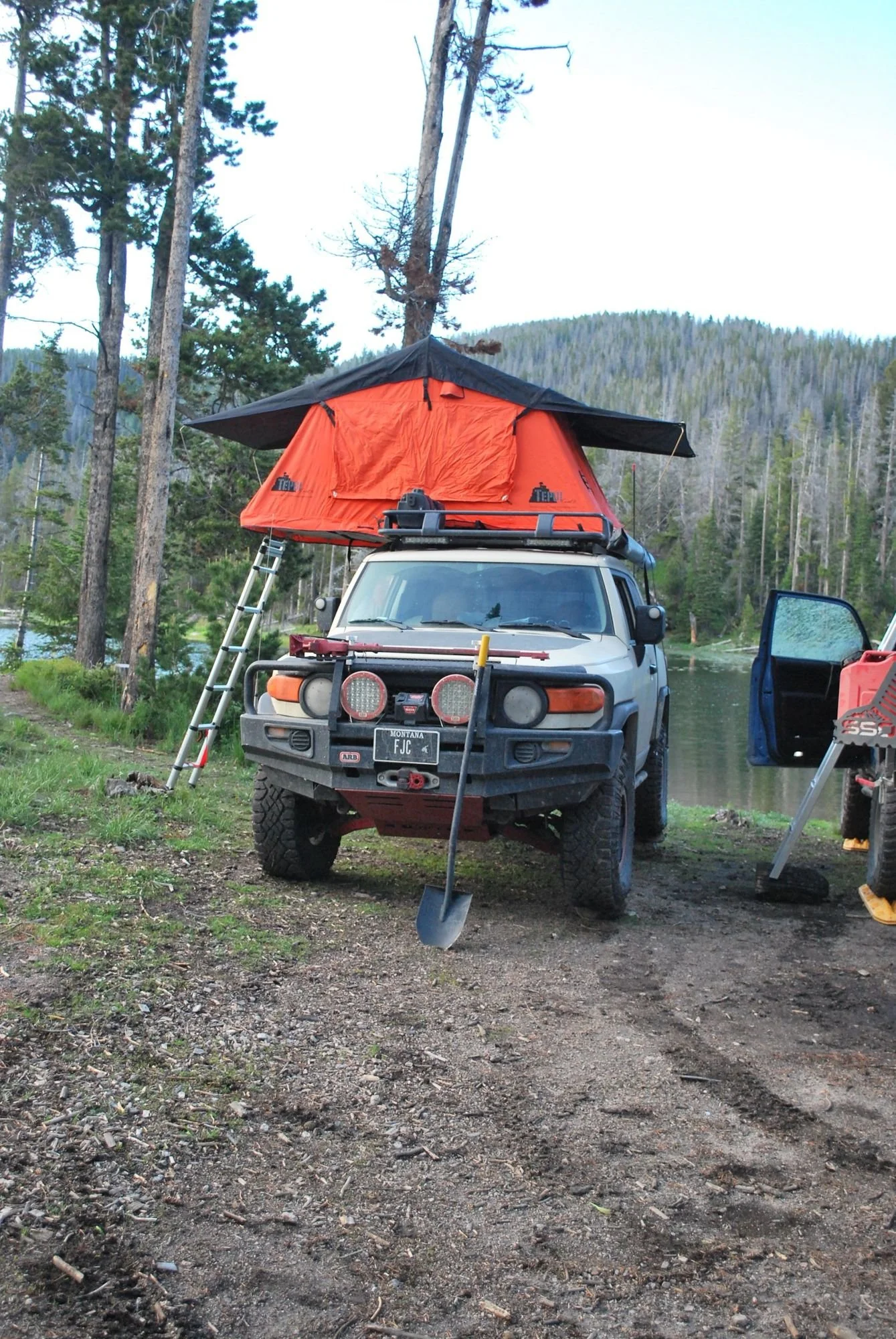 Tan FJ Cruiser with an open orange rooftop tent and ladder parked near a lake in the pines on the North Meadow Creek Twin Lakes trail in the Tobacco Root Mountains, Montana