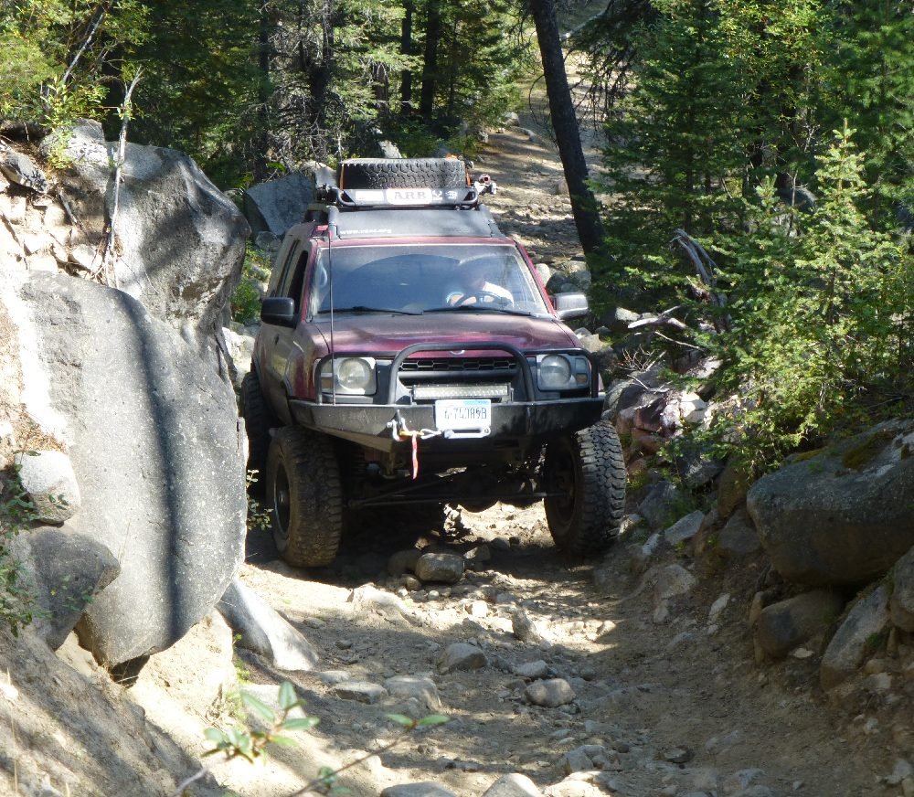 Red Nissan Xterra driving through a narrow gap between large boulders on a forest track