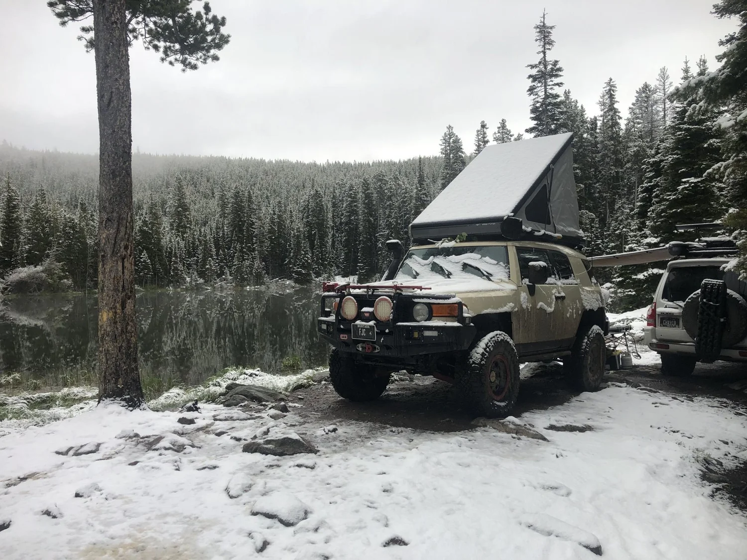 Tan FJ Cruiser with a hard-shell rooftop tent at a snowy lakeside camp in the pines in the Elkhorn Mountains near Boulder, Montana