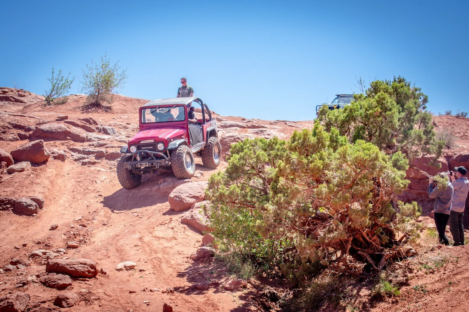 Red caged Toyota FJ40 climbing a rocky ledge while people watch on the Seven Mile Rim trail near Moab, Utah