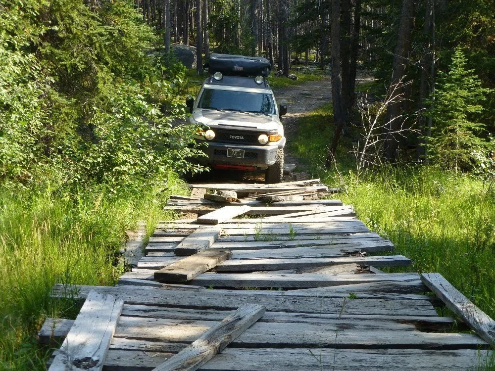 Tan FJ Cruiser crossing an old wooden plank bridge over a grassy creek on a rocky forest trail in Montana