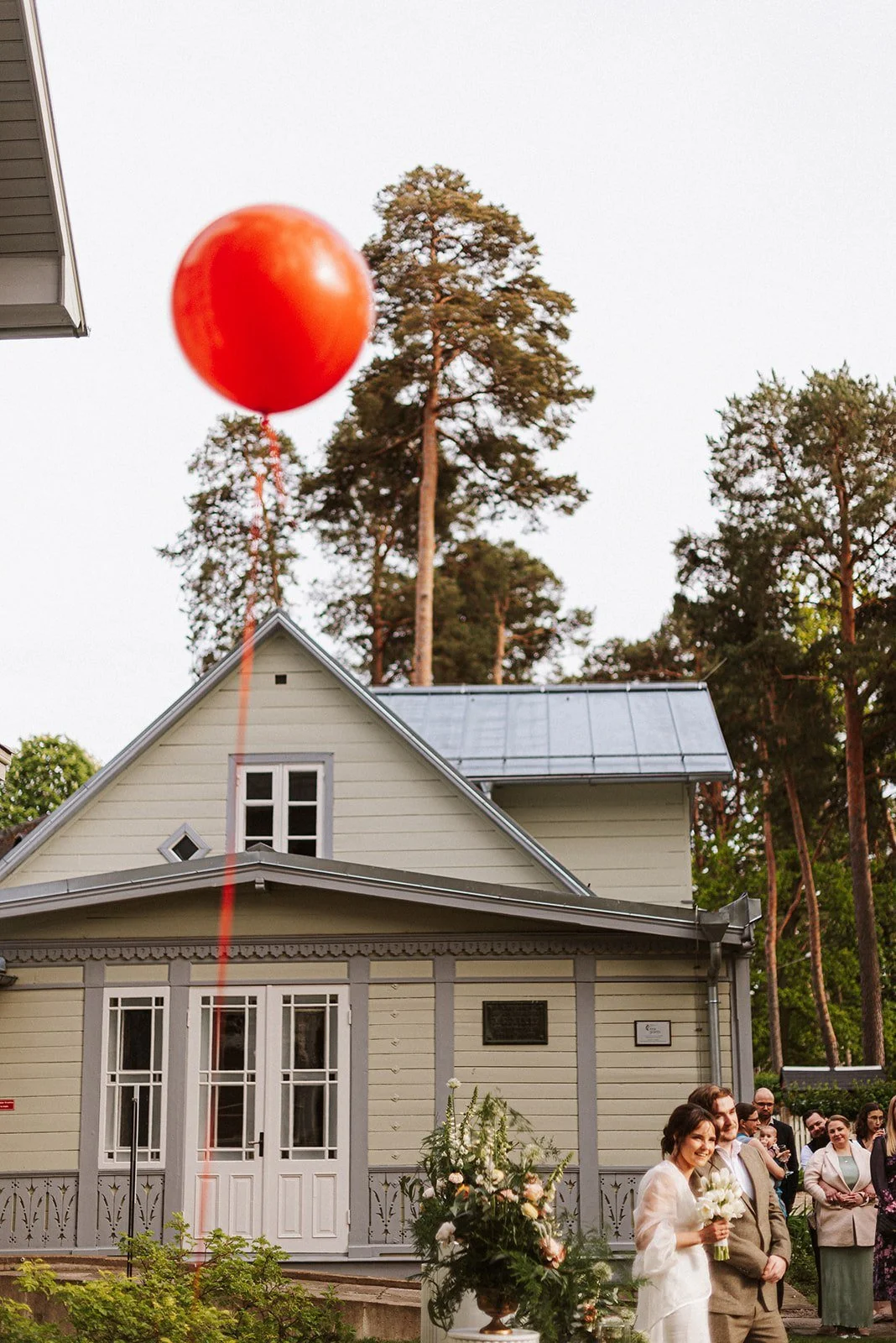 Sarkans balons paceļas virs cilvēkiem pie koka ēkas.