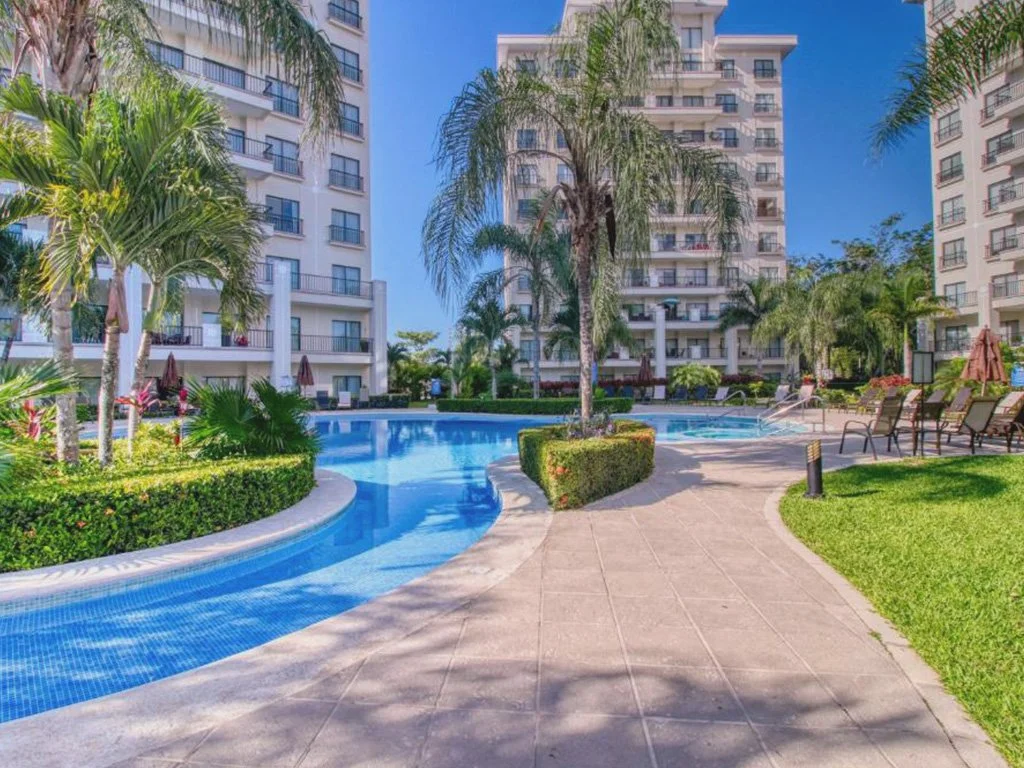 Resort-style swimming pool at Jacó Bay condominium in Jacó, Costa Rica, surrounded by palm trees and modern residential towers.
