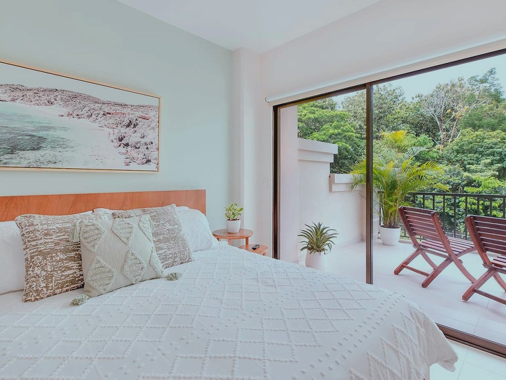 Bright master bedroom at Jacó Bay condominium with sliding glass doors leading to a private balcony overlooking tropical greenery.