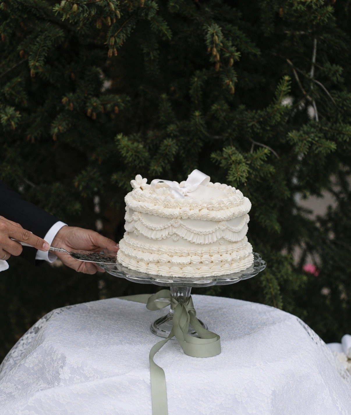 A ruffled wedding cake on a glass stand tied with green ribbon
