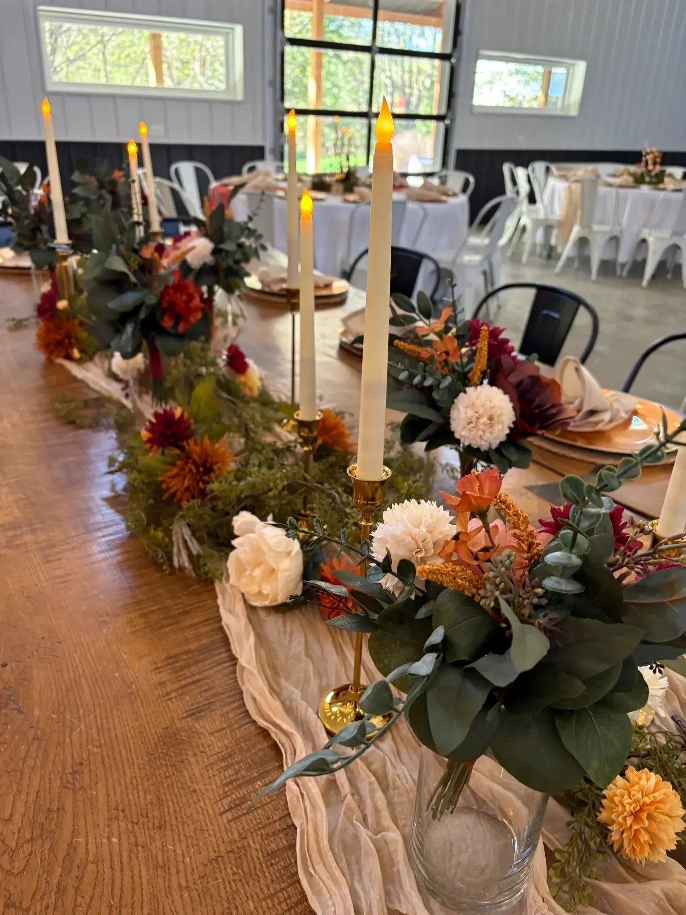 Long floral table centerpiece in warm autumn colors with tall lit candles, arranged along a dining table in a bright room.