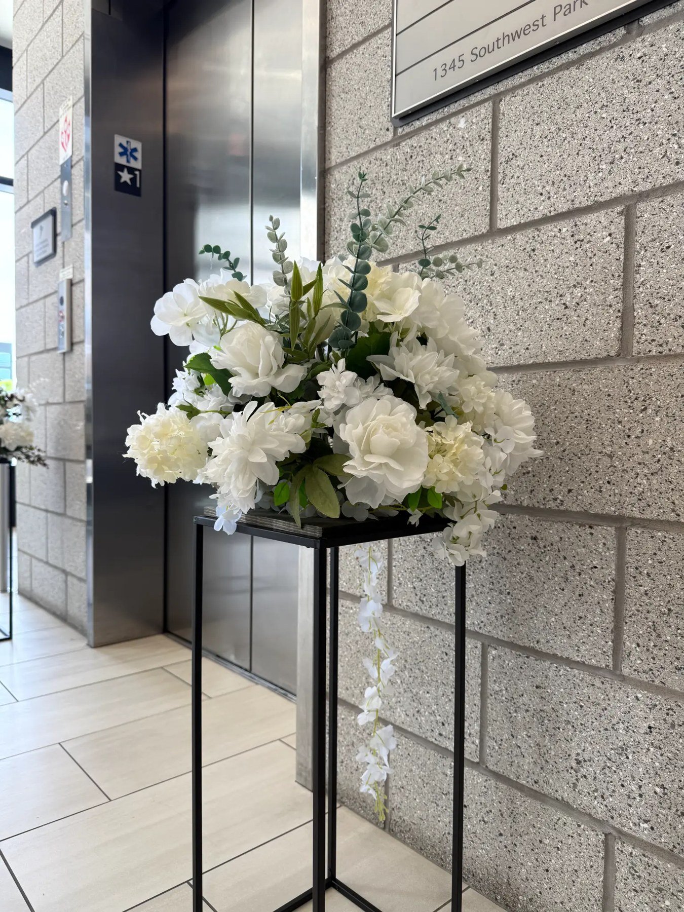 White and ivory floral arrangement with eucalyptus and trailing flowers on a tall black pedestal.