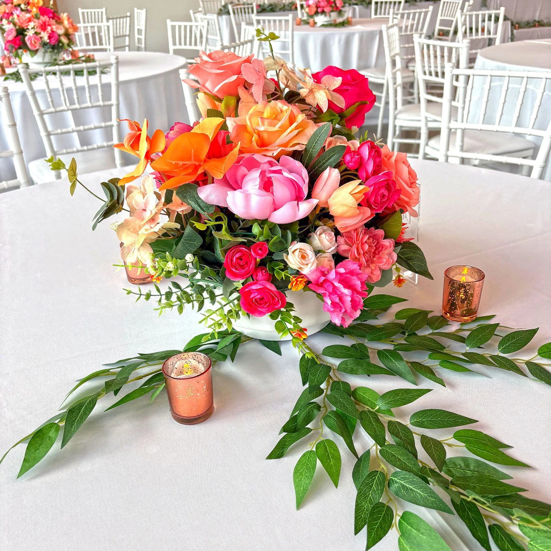 Colorful pink, coral, orange, and peach flower centerpiece with trailing greenery and votive candles on a white banquet table.