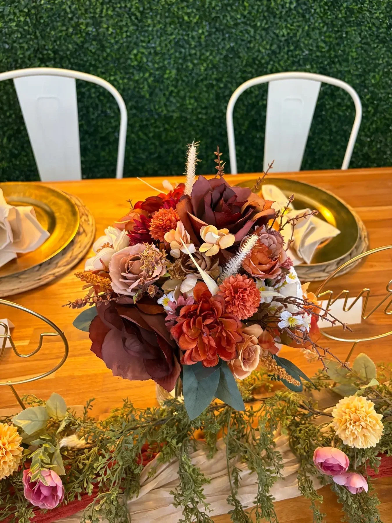 Colorful burgundy, rust, peach, cream, and mauve floral centerpiece on a dining table with gold place settings and trailing greenery.
