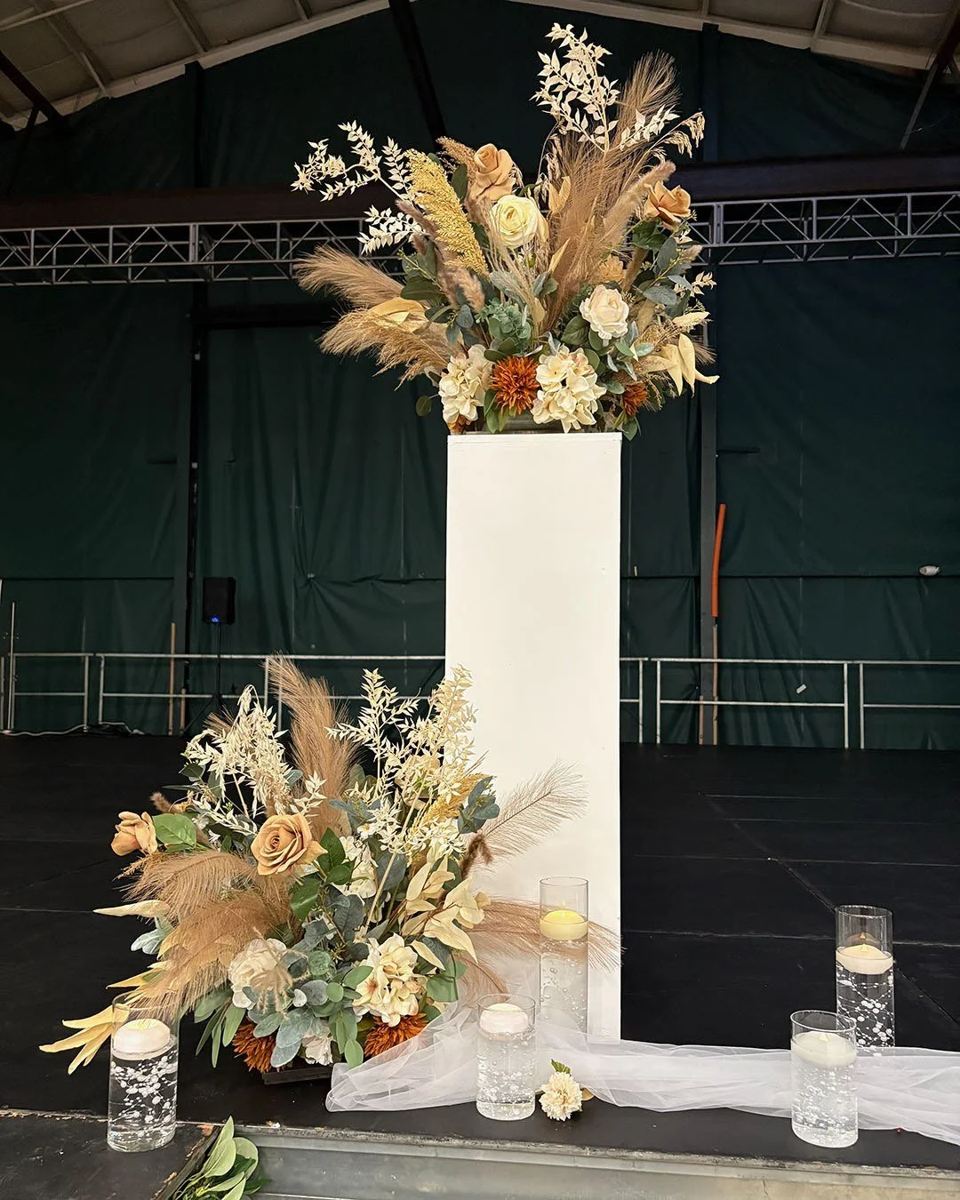 Floral ceremony-style installation with two cream-and-peach arrangements on and beside a tall white pedestal, surrounded by candles and sheer fabric on a dark stage.