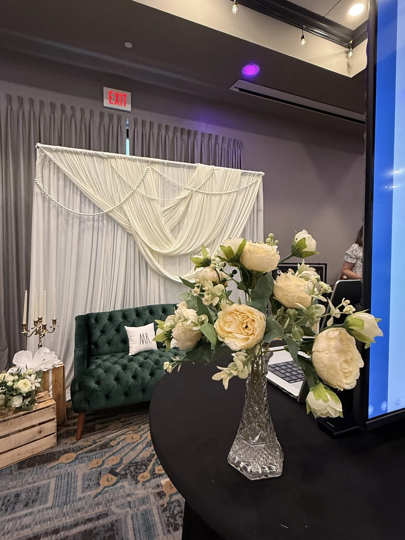 Cream and white flower centerpiece in a faceted glass vase on a black table, photographed in front of a draped ceremony backdrop and dark green sofa.