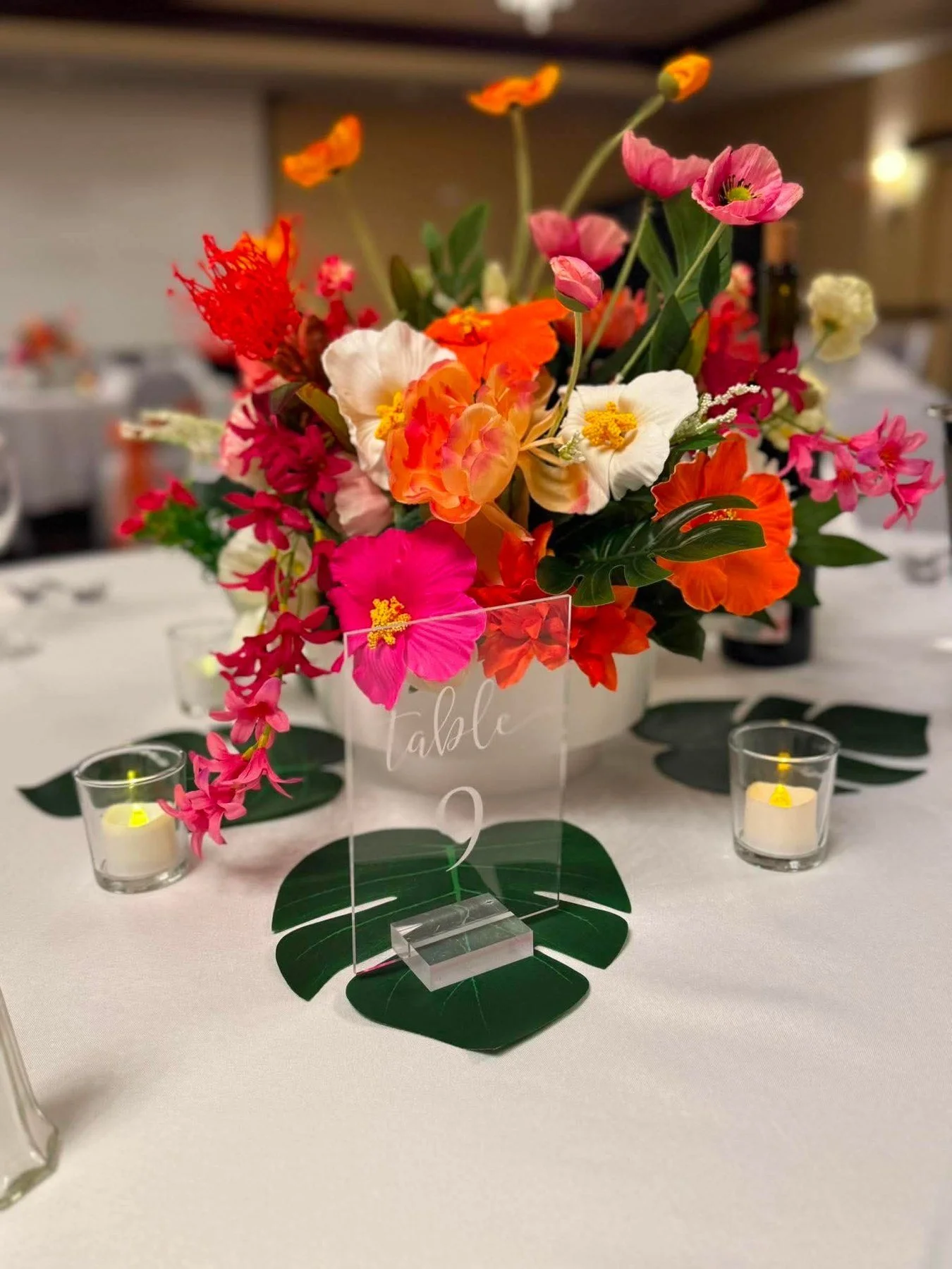 Colorful pink, orange, red, and white floral centerpiece with green leaves, votive candles, and a clear table-number sign on a banquet table.