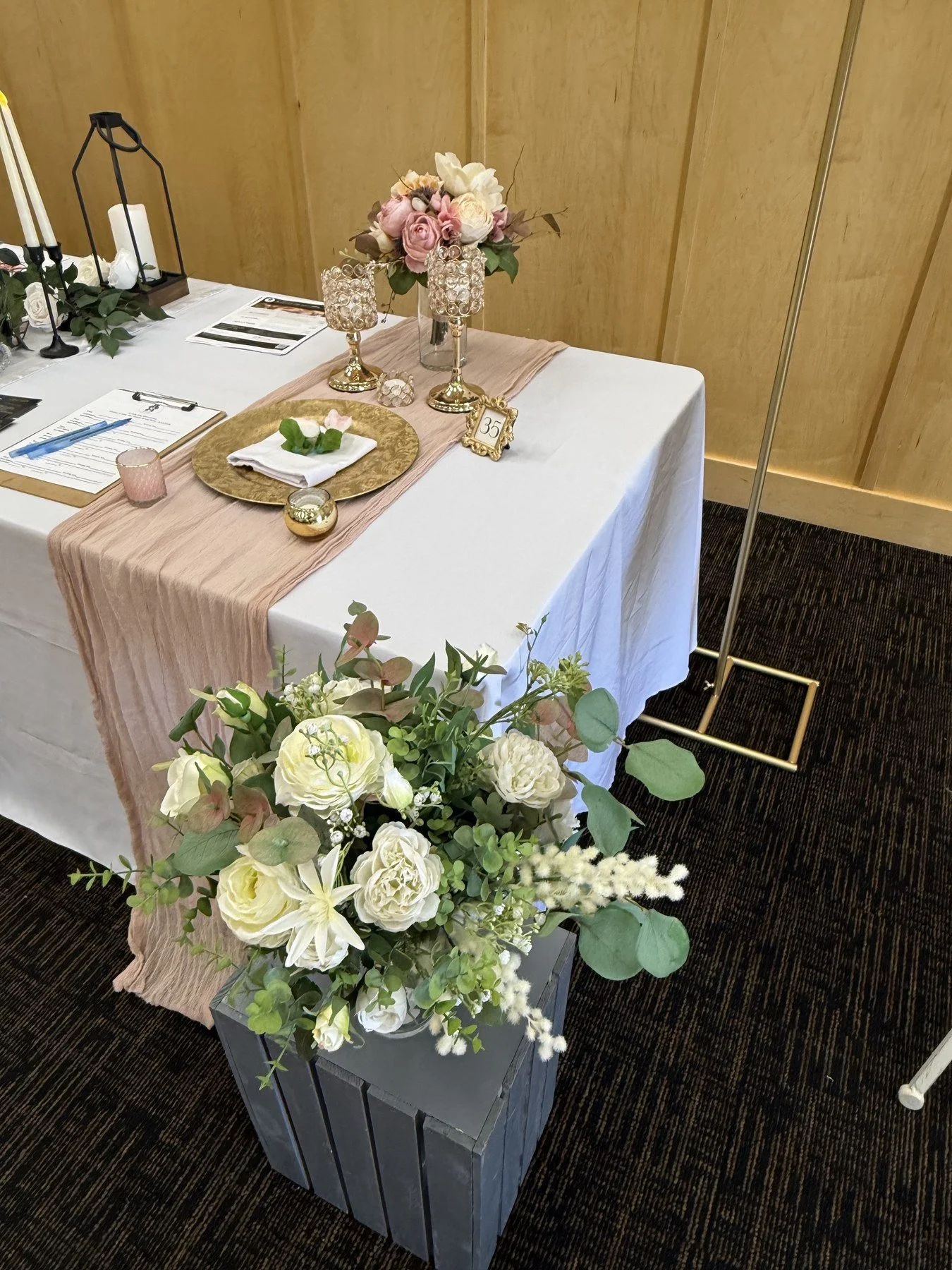 Large white and cream floral arrangement with green foliage on a dark pedestal in front of a decorated blush-and-white table with candles and a smaller pink floral arrangement.