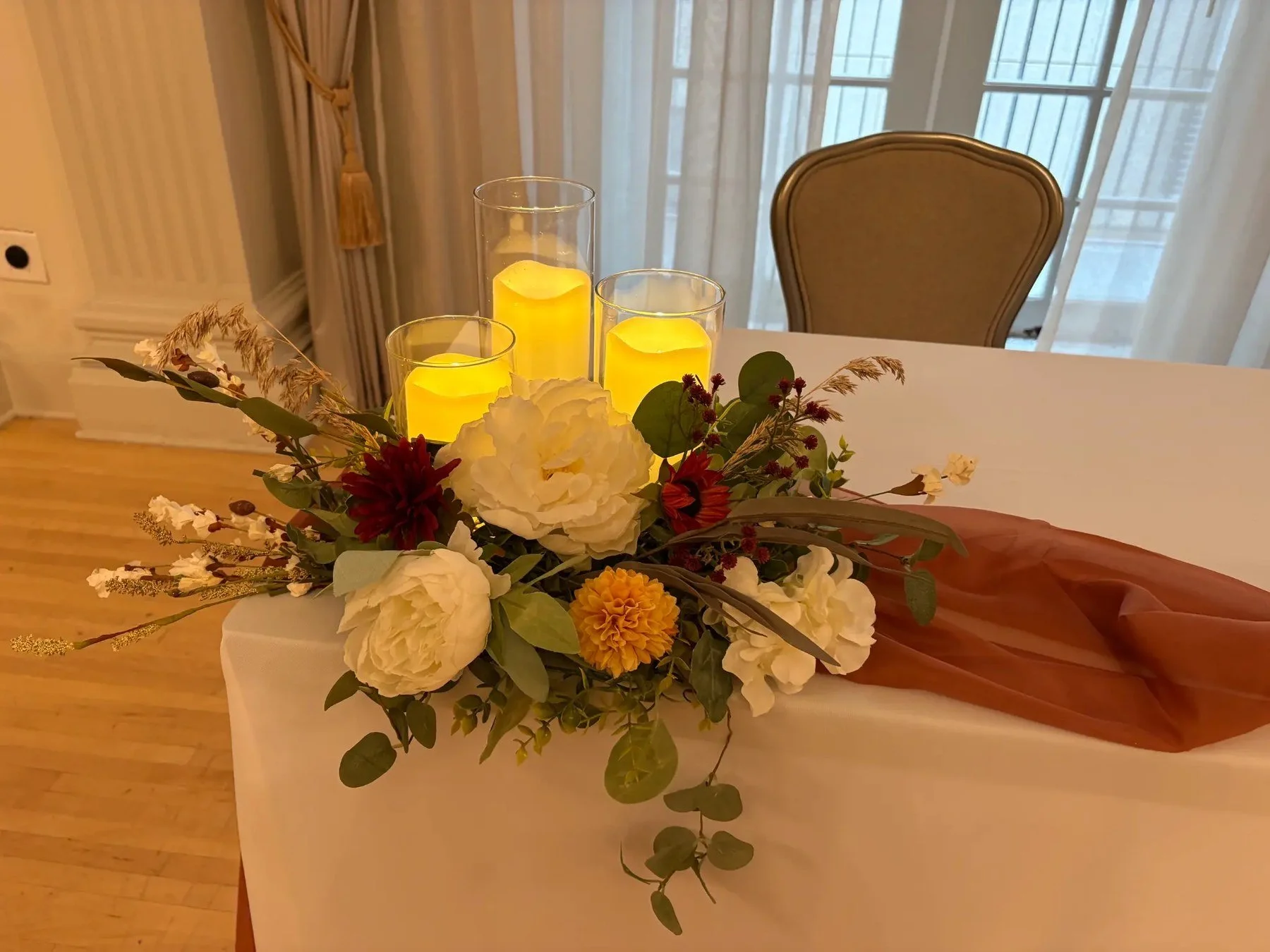 Wide floral centerpiece with cream, white, red, and yellow flowers, greenery, and three glowing candles on a draped table in a warm room.