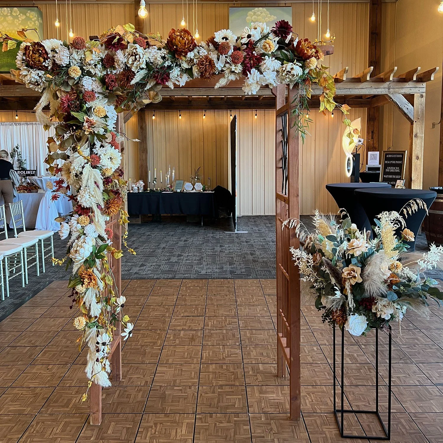 Floral arch covered in cream, white, burgundy, and golden blooms inside a rustic event venue, with a separate flower stand at lower right.