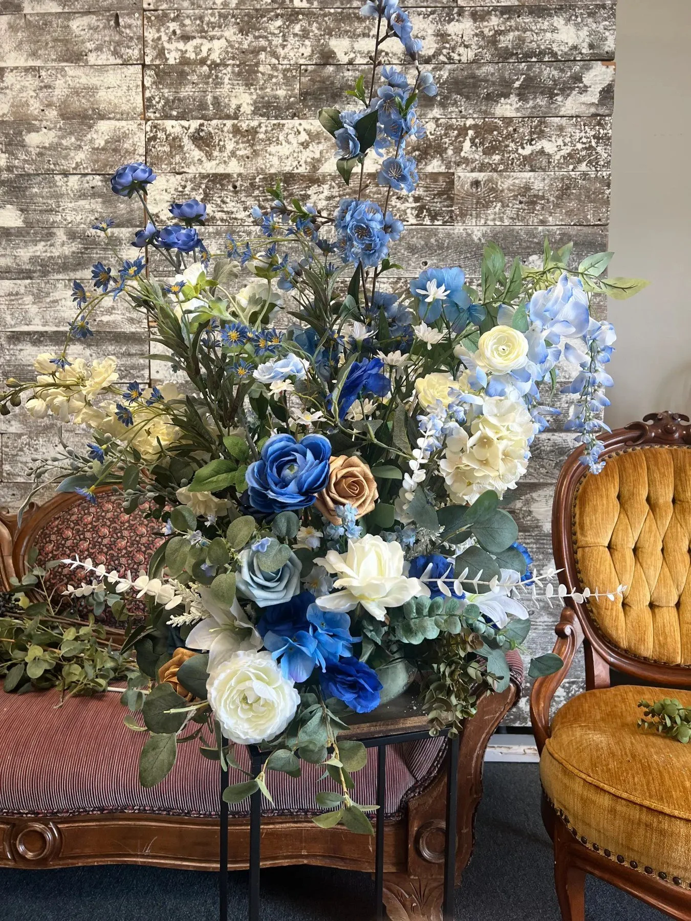 Blue, ivory and cream floral arrangement with greenery on a tall black pedestal in front of a weathered wood backdrop.