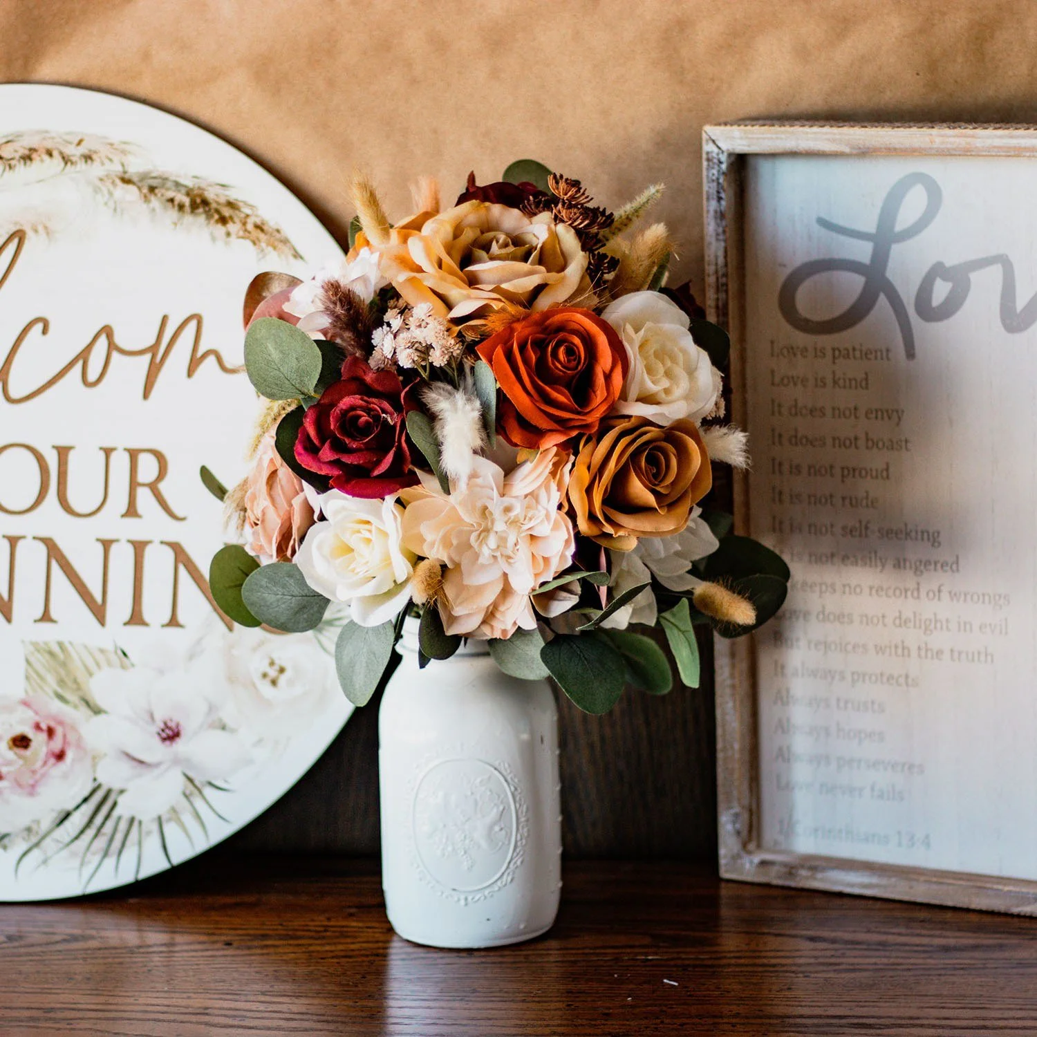 Colorful floral centerpiece in a white vase on a wooden table, arranged between a decorative plate and a framed quote print.