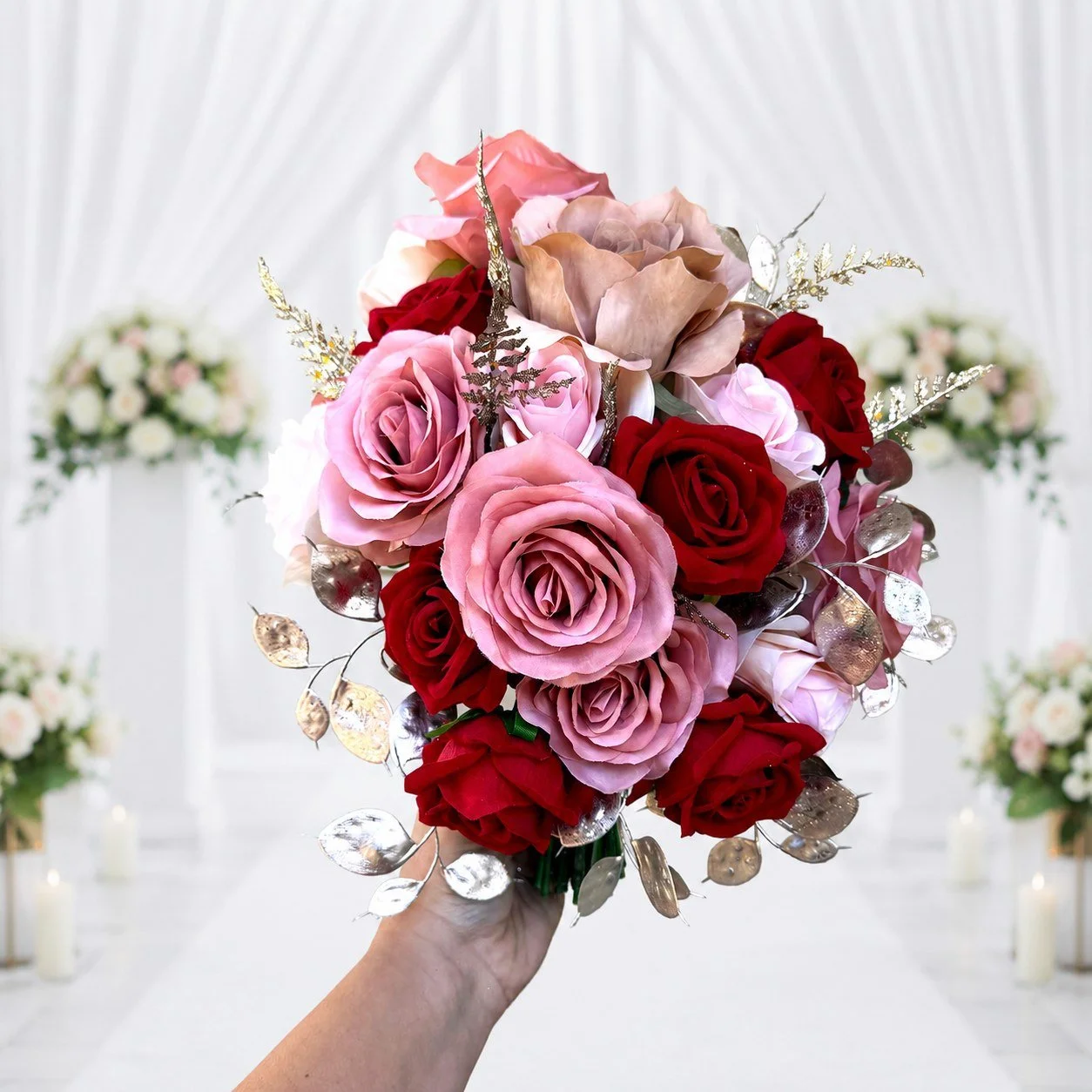Hand holding a rounded pink and deep red floral bouquet with metallic leaf accents against a bright white ceremony backdrop.