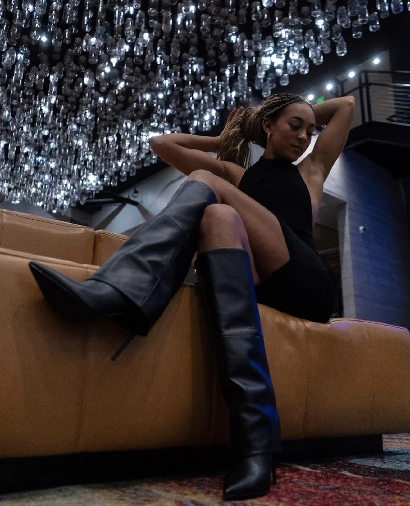Woman in a black outfit and tall boots reclining on a sofa beneath decorative lights.