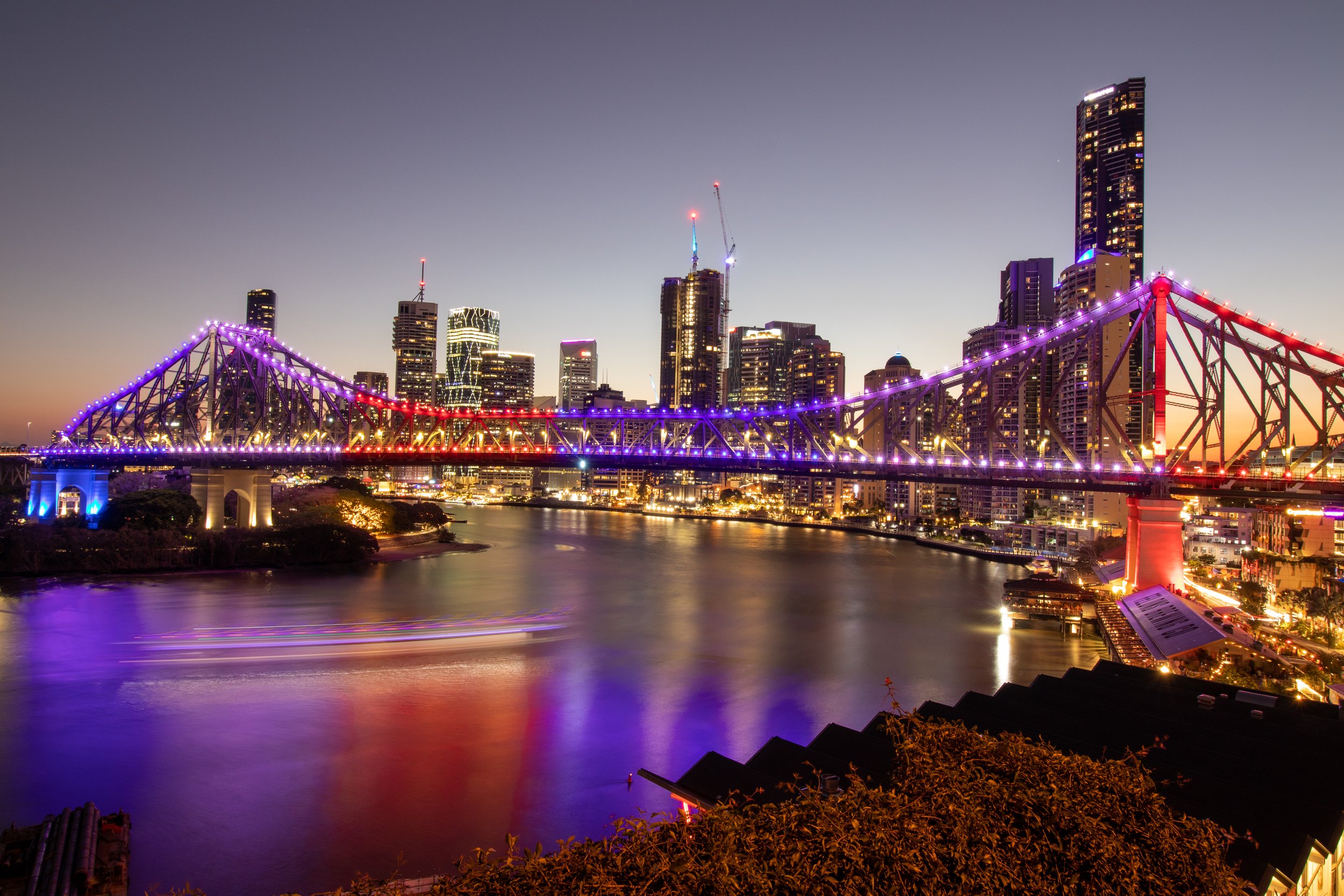 Brisbane's city skyline and the Story Bridge lit up at sunset, with a ferry moving along the river below