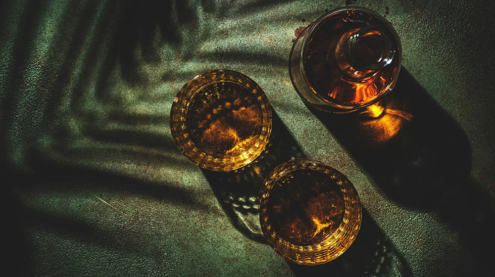 Top-down view of a whiskey decanter and two glasses casting dramatic shadows in a dimly lit speakeasy setting.