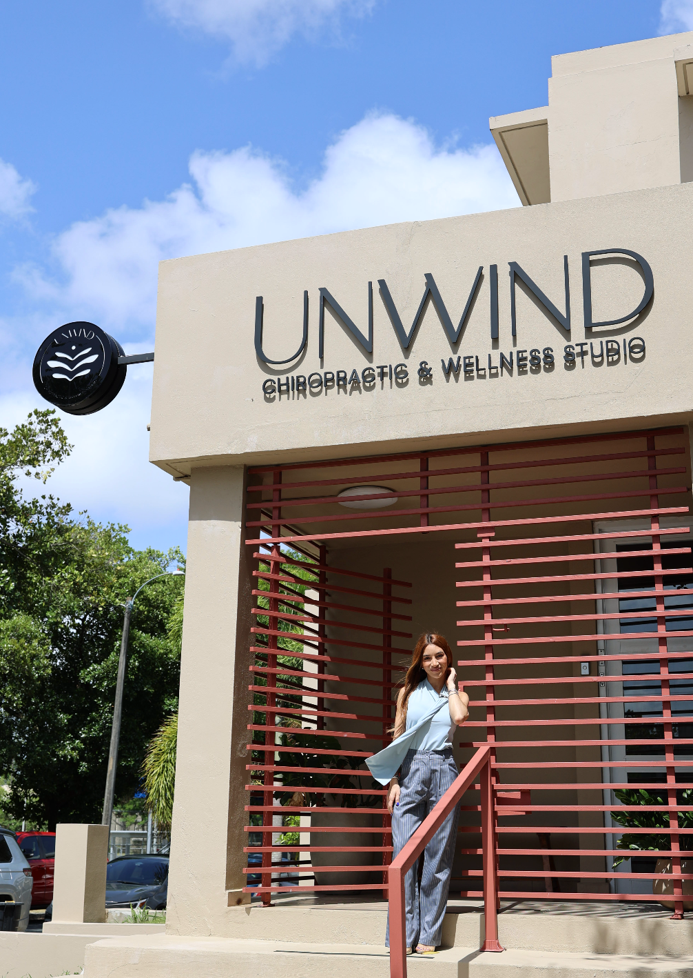 Una mujer de pie en la entrada de Unwind Chiropractic & Wellness Studio, bajo el rótulo del estudio y el cielo azul