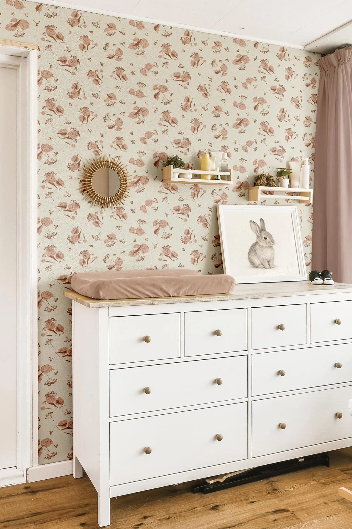 Blush pink botanical nursery wallpaper featuring hand-drawn autumn leaves and delicate flowers on a warm cream background in a boho baby changing area.