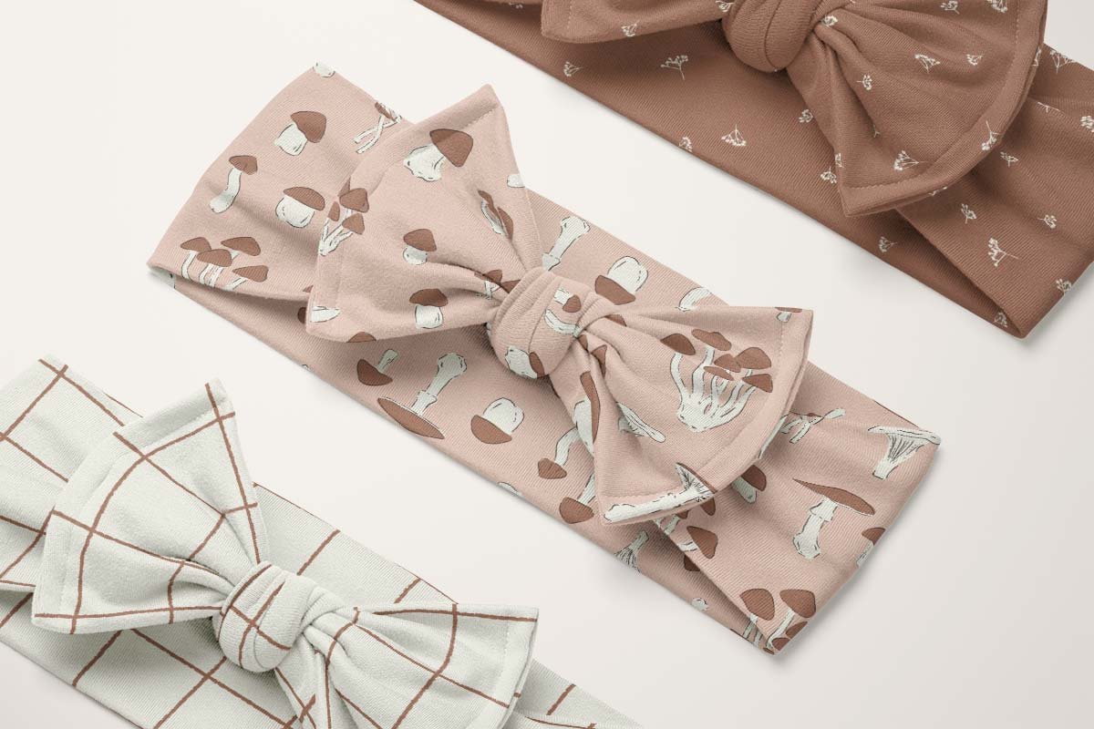 Earthy boho fall fabric patterns featuring hand-drawn mushrooms, botanical sprigs and checks, shown on soft bow headbands for babies and children.