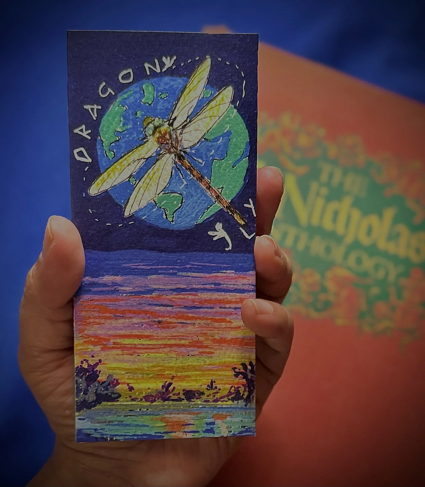 Hand holding the N'marks Dragonfly Bookmark in front of a vintage book