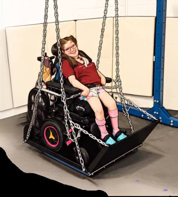 A girl smiles while sitting in her wheelchair, secured inside an adaptive wheelchair swing.