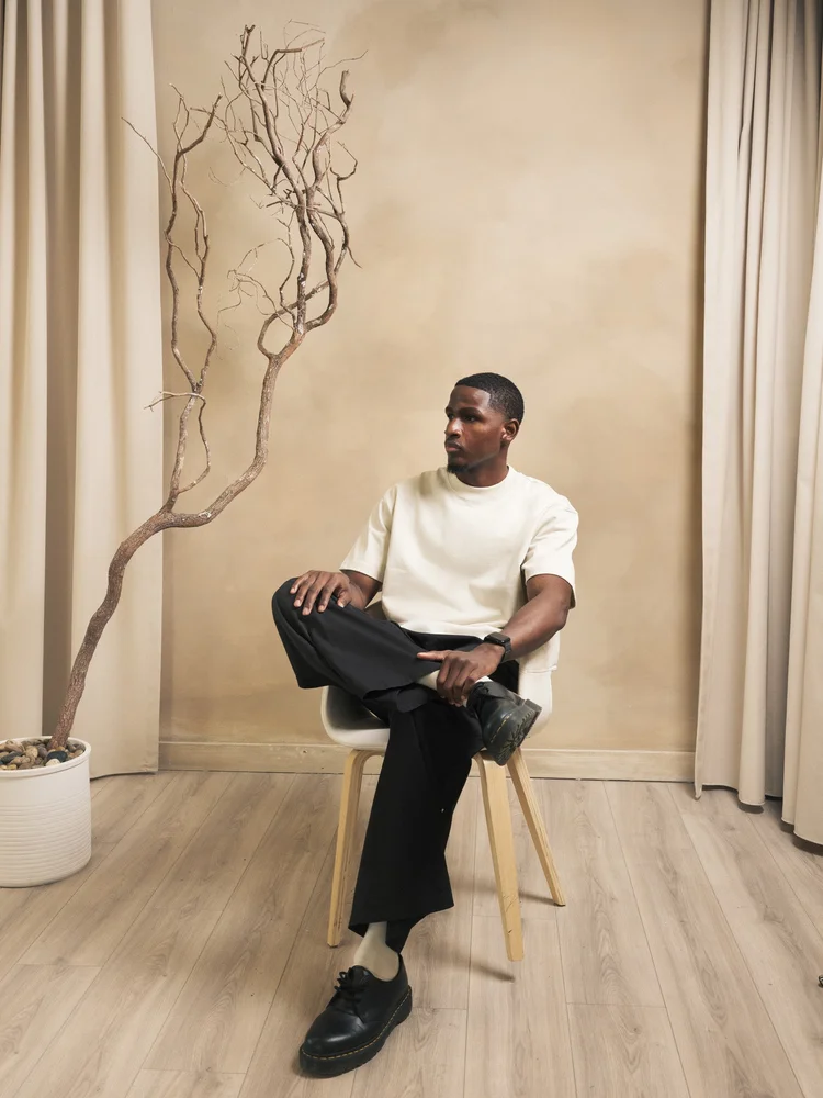 Seated model wearing a cream R3 CONCEPT T-shirt beside a sculptural tree