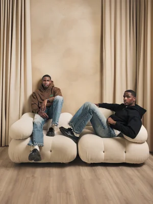 Two models wearing brown and black R3 CONCEPT hoodies on a sculptural cream sofa