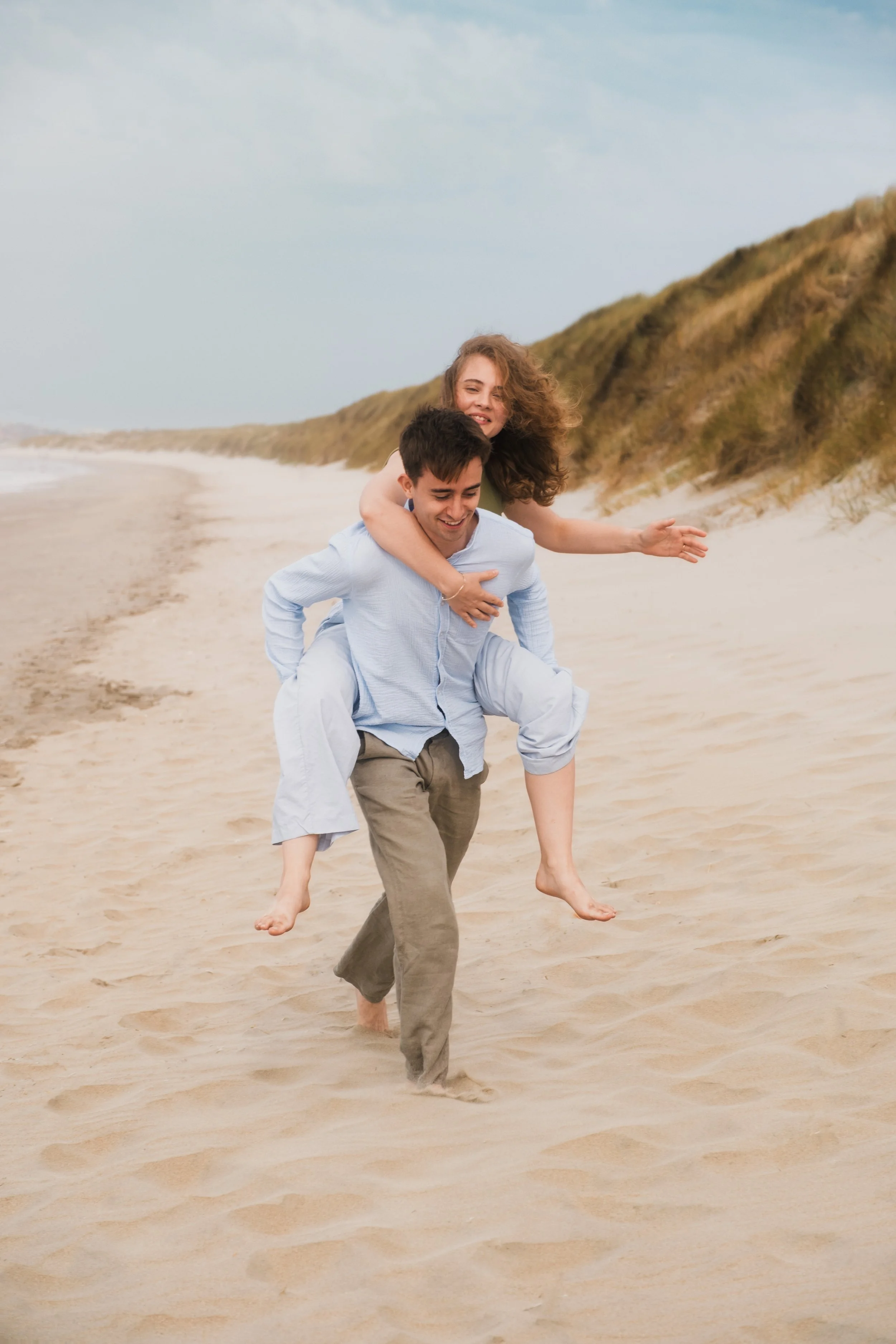 Couple joyeux sur la plage, l'un porte l'autre sur son dos, séance photo couple en Belgique, Marion David photographe
