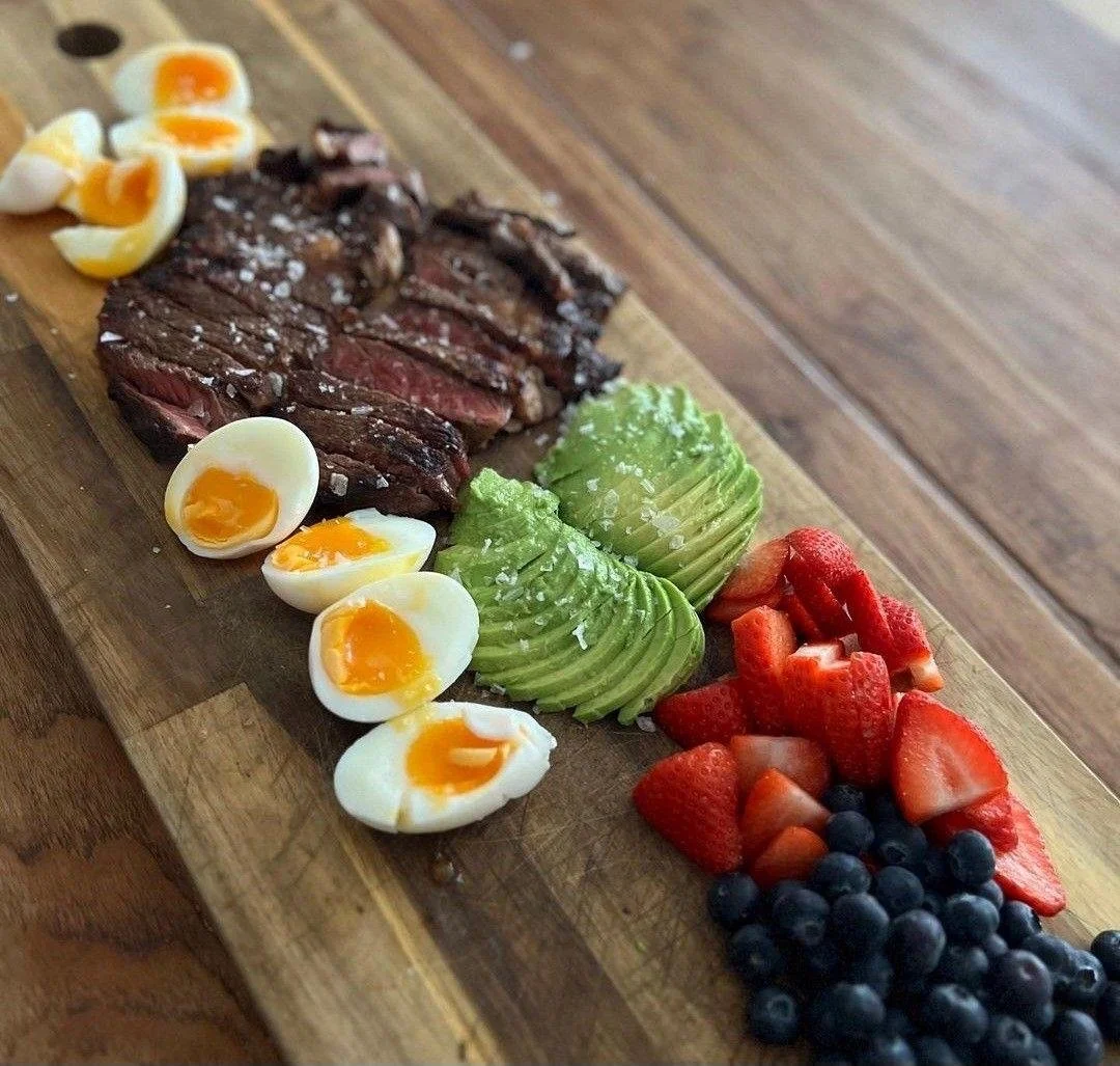 Sliced steak, boiled eggs, avocado, strawberries, and blueberries on a wooden serving board