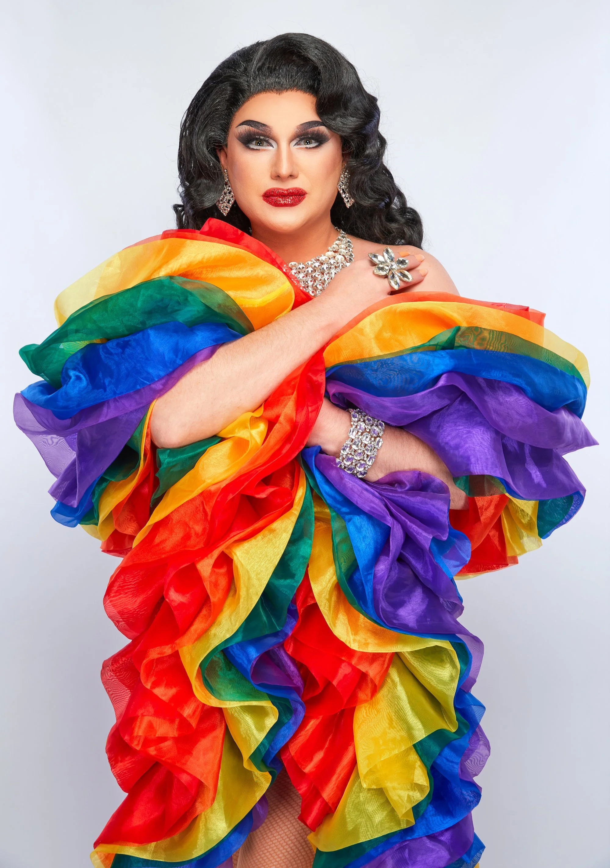 Charisma Belle with dark waves, sparkling jewellery and a vibrant rainbow ruffled outfit