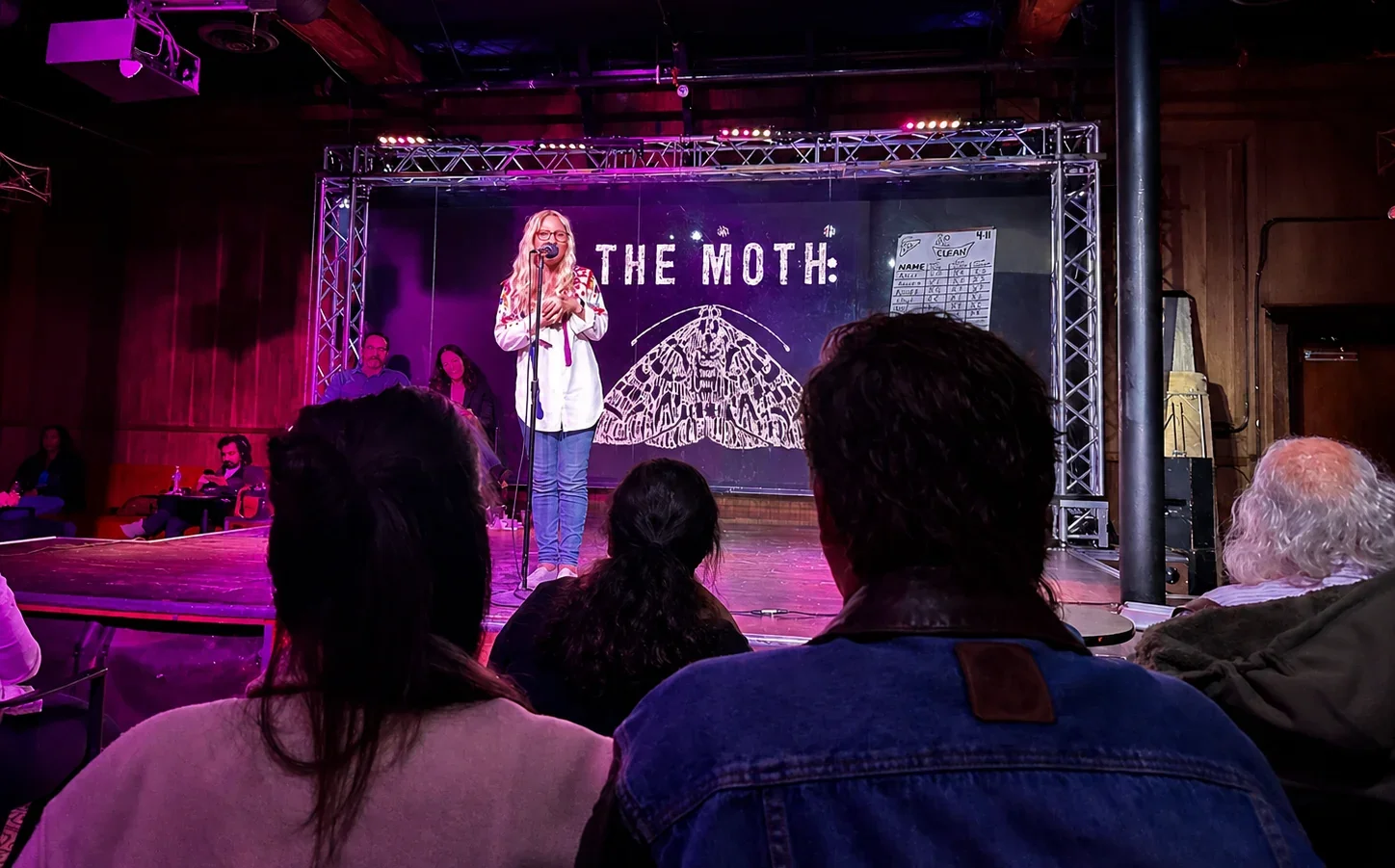 Ali Blackmon speaking at a microphone beneath The Moth sign, viewed from the seated audience.