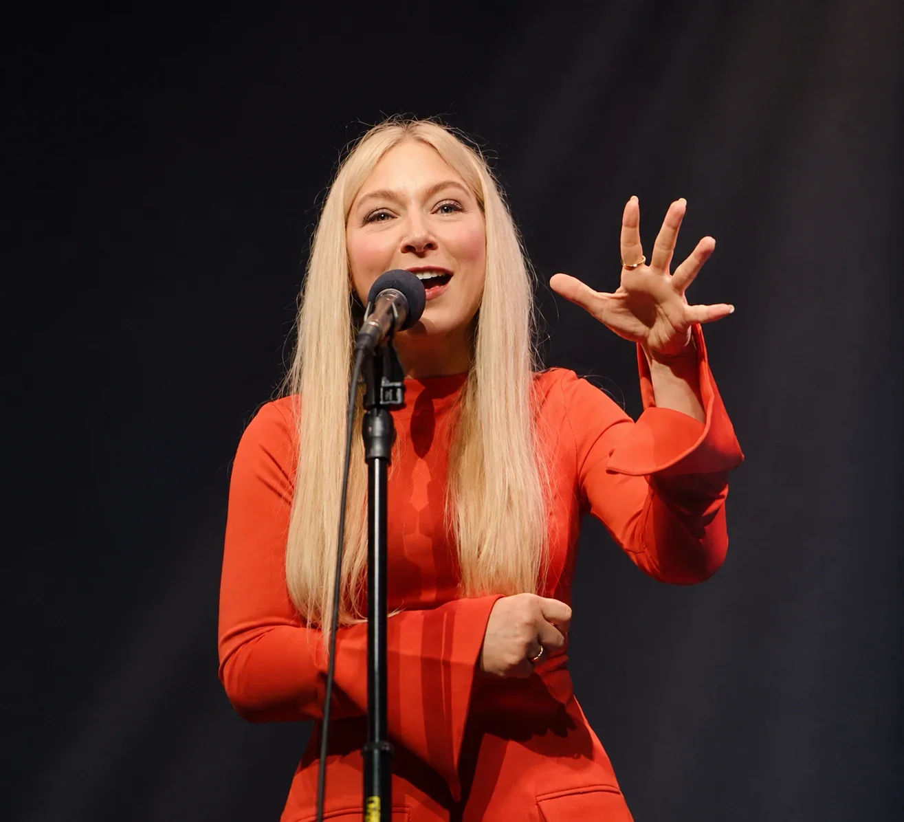 Ali Blackmon in red, speaking into a microphone with one hand raised.