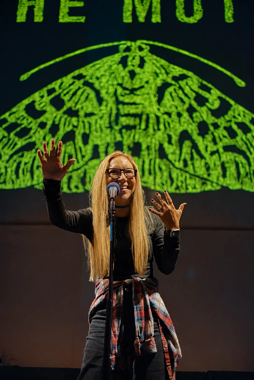 Ali Blackmon gesturing at a microphone beneath a green moth backdrop.