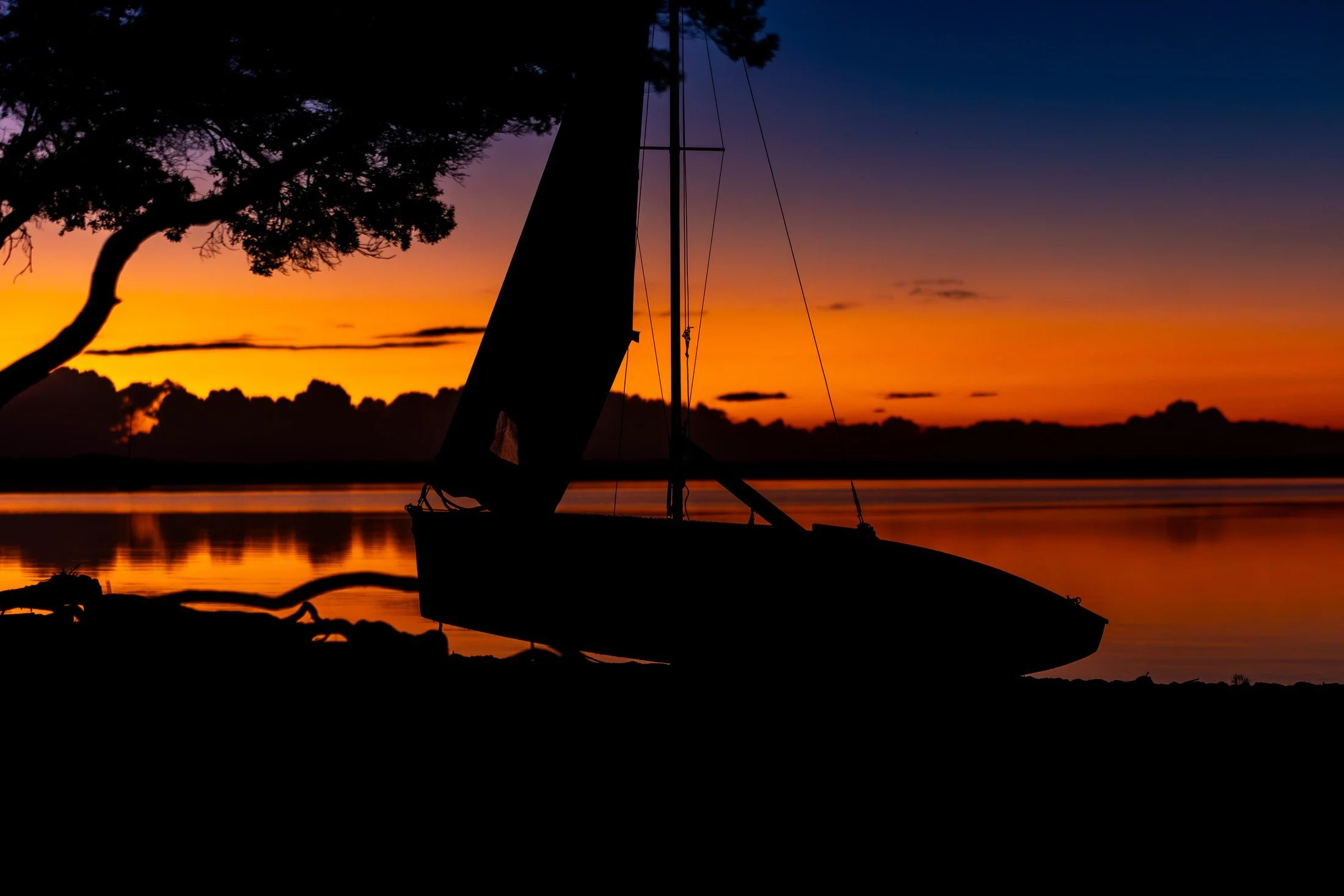 A silhouetted sailboat and trees beside still water under a deep blue and orange sunset.