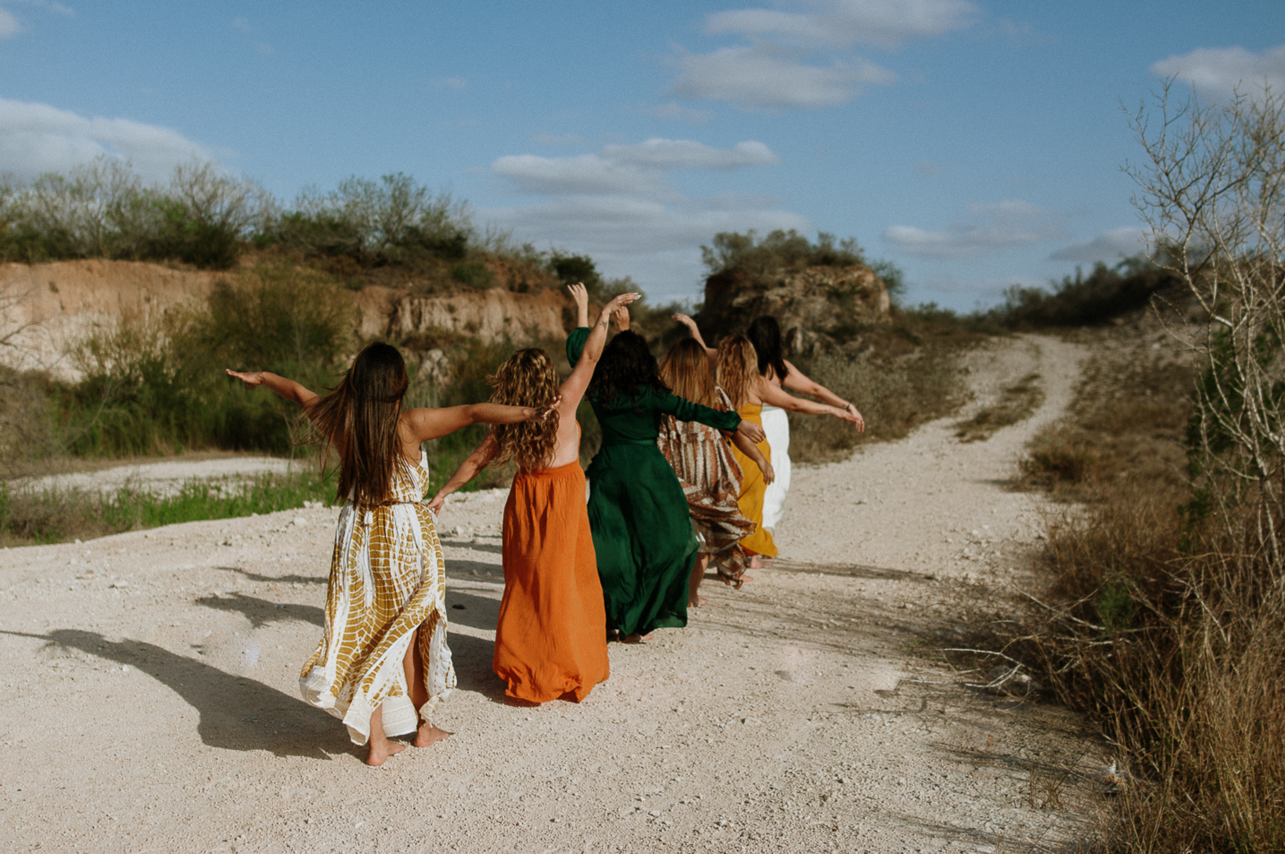 Seven women in colorful dresses walking together down a sunlit dirt road, arms raised in celebration