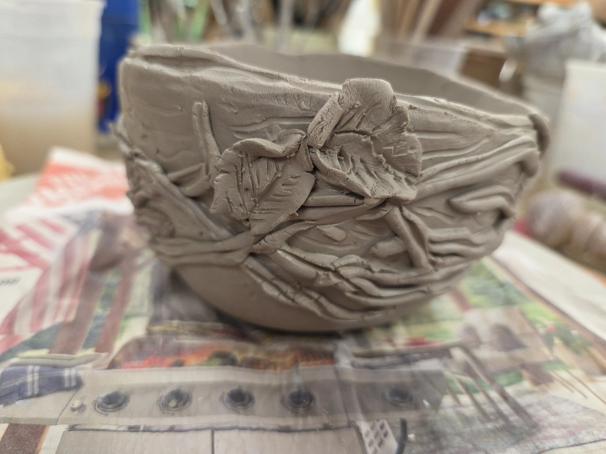 Unfired handbuilt vessel by Moe Lewis-Wolf with an undulating rim, layered bands, and applied leaves