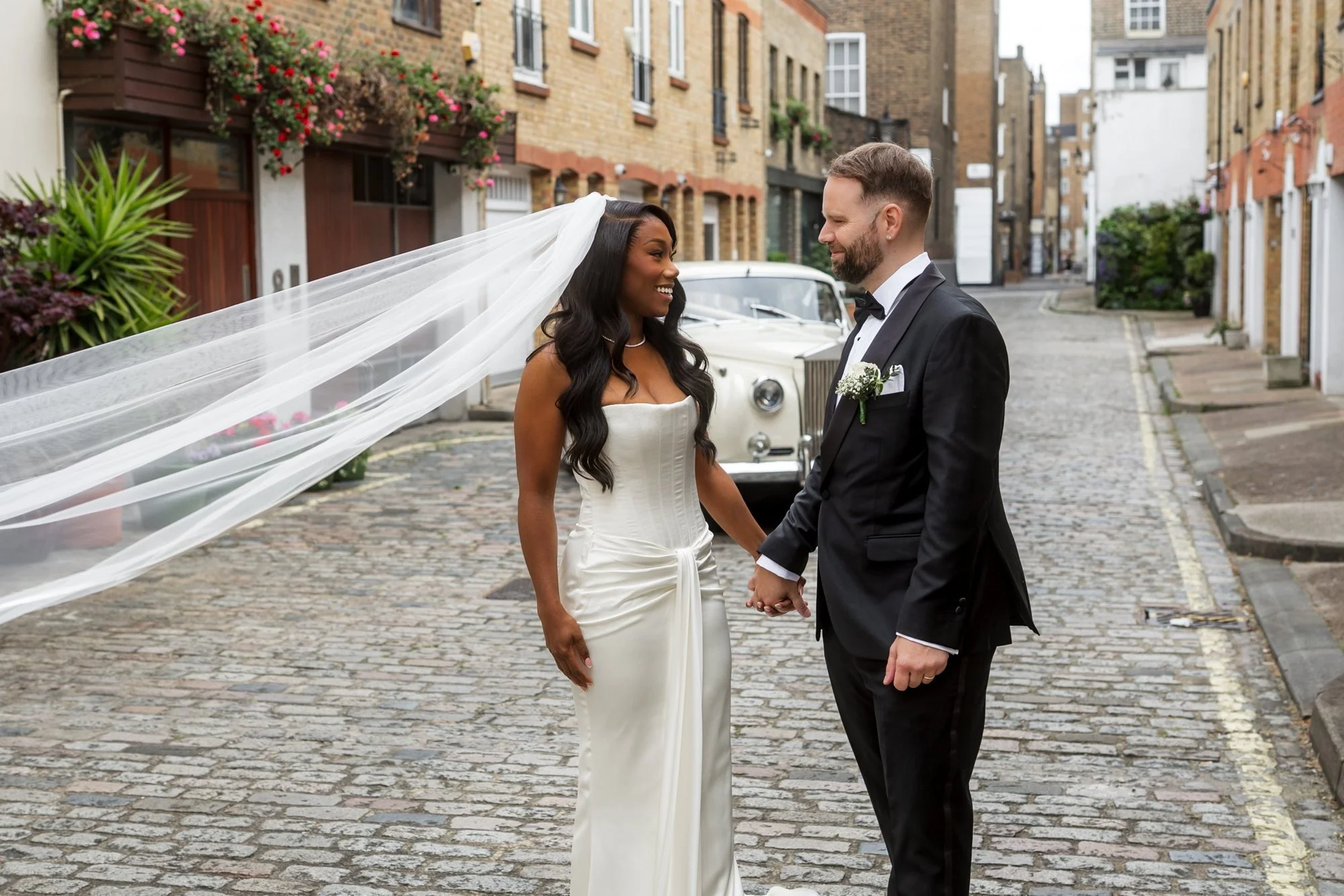 Bride and groom hand in hand in a London mews, her veil lifted by the wind, by London wedding photographer Vino Captures