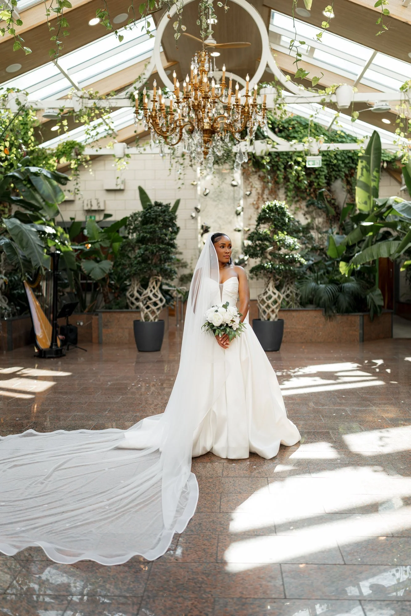 Bride beneath a crystal chandelier in a glasshouse wedding venue, her veil flowing across the floor, by London wedding photographer Vino Captures