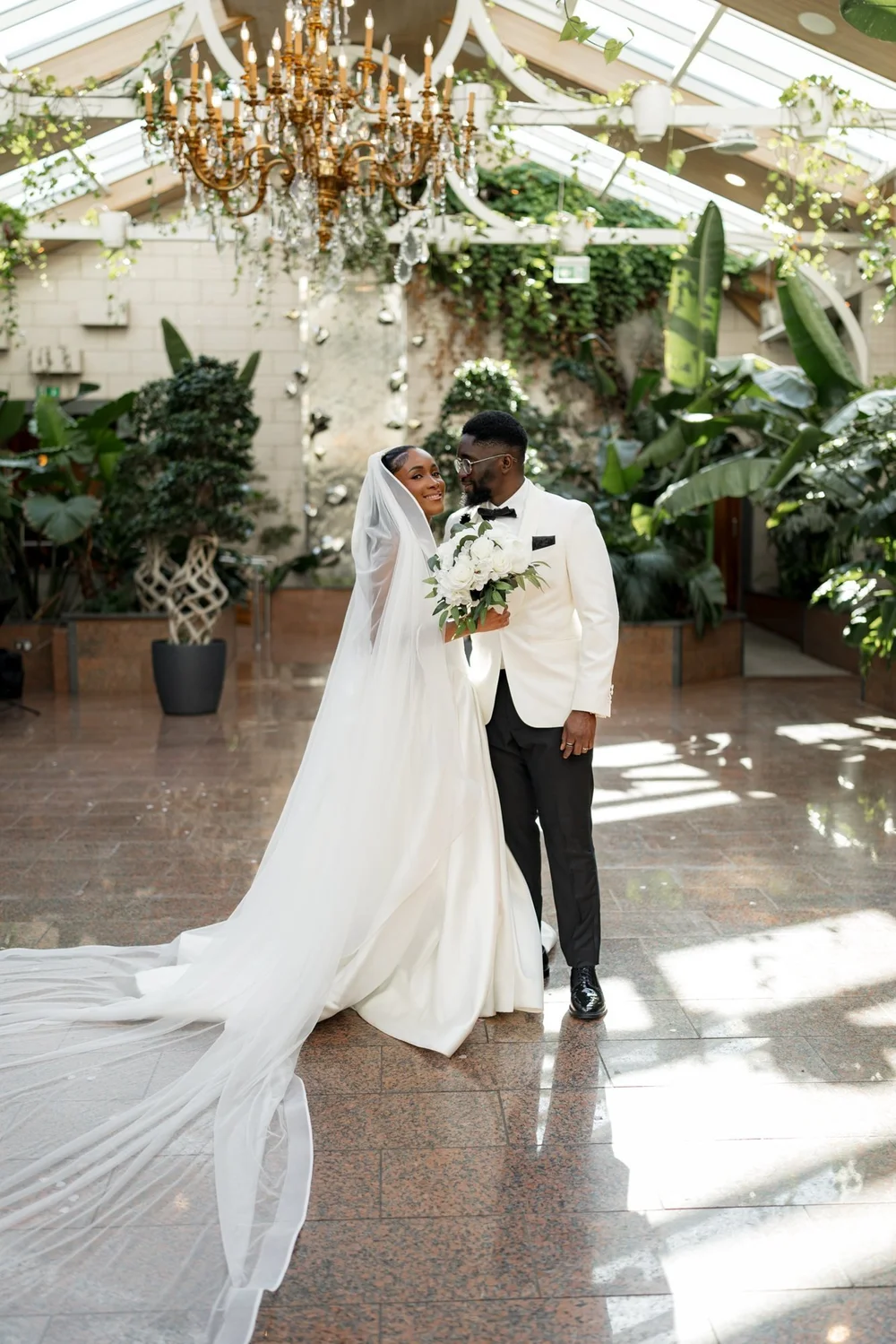 Bride and groom posed beneath a chandelier in a glasshouse venue