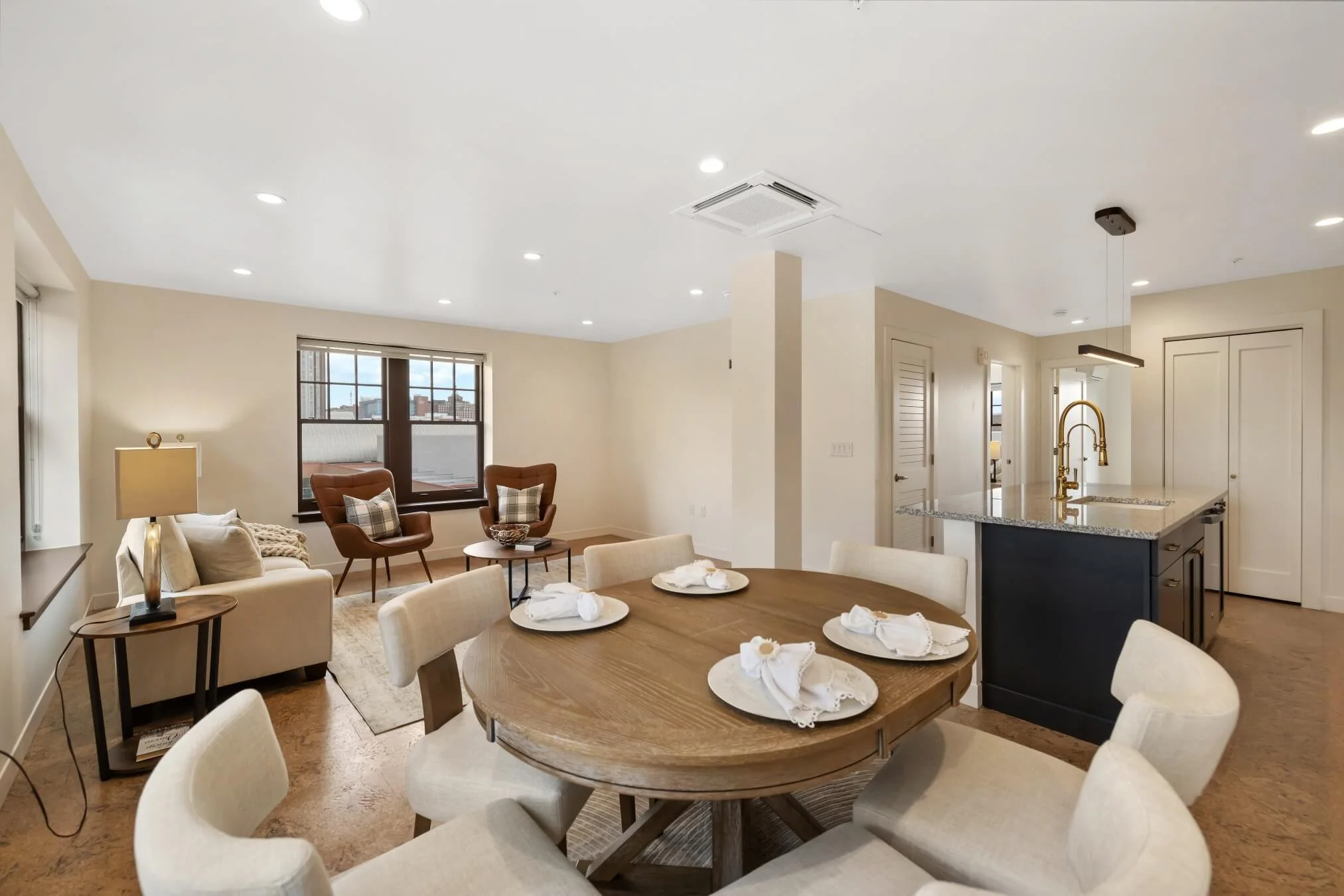Open concept living and dining area with round wood table upholstered chairs leather accent chairs and large windows providing natural light